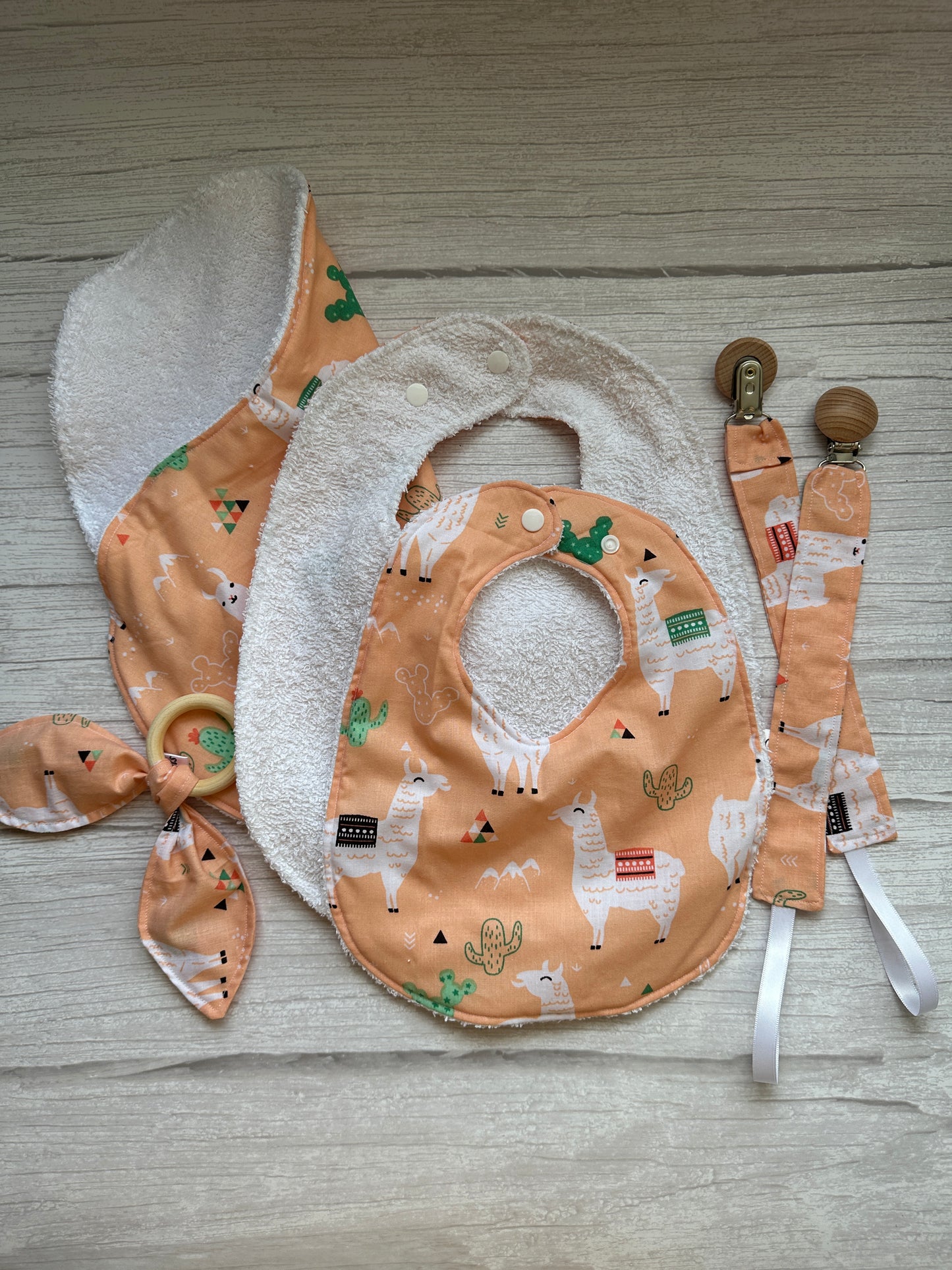 The Cotton & Terry Towelling Baby Sets – Llama Loves by Crafts by Kate features a handcrafted cotton collection in peach and white, with charming llama and cactus designs. Includes an ultra-soft bib, pacifier clip, burp cloth, and a knotted headband elegantly displayed on light wood.