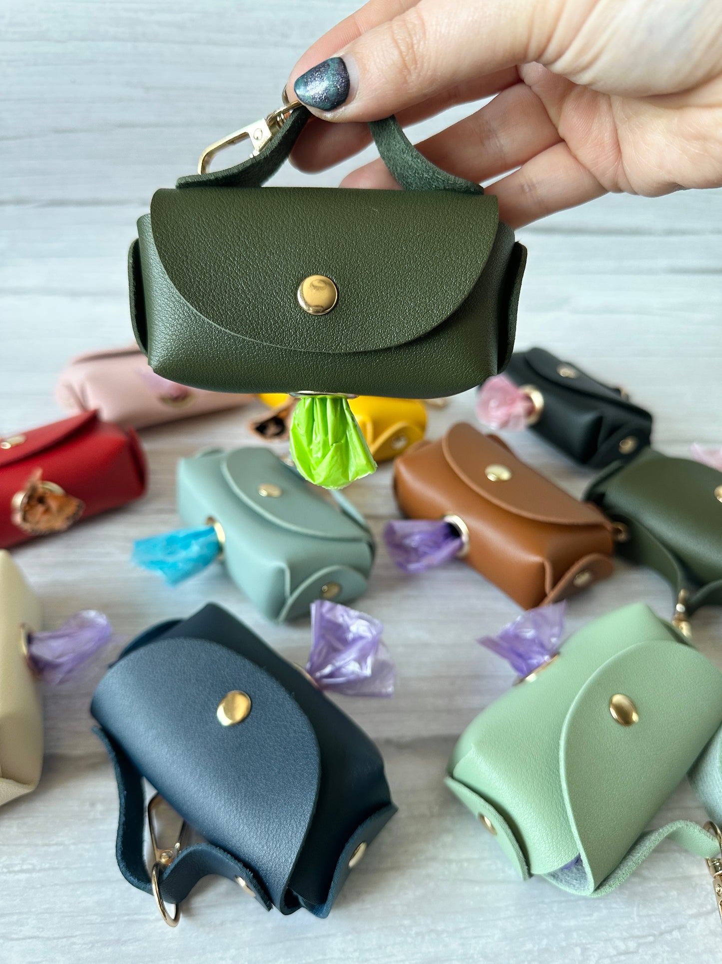A hand displays a small, handbag-shaped green leather pouch with a metal clasp, identified as Crafts by Kate's Pet Poo Bag Holder Handbag. In the background, various faux leather pouches in different colors rest on light wood, each showing a partially visible waste bag.