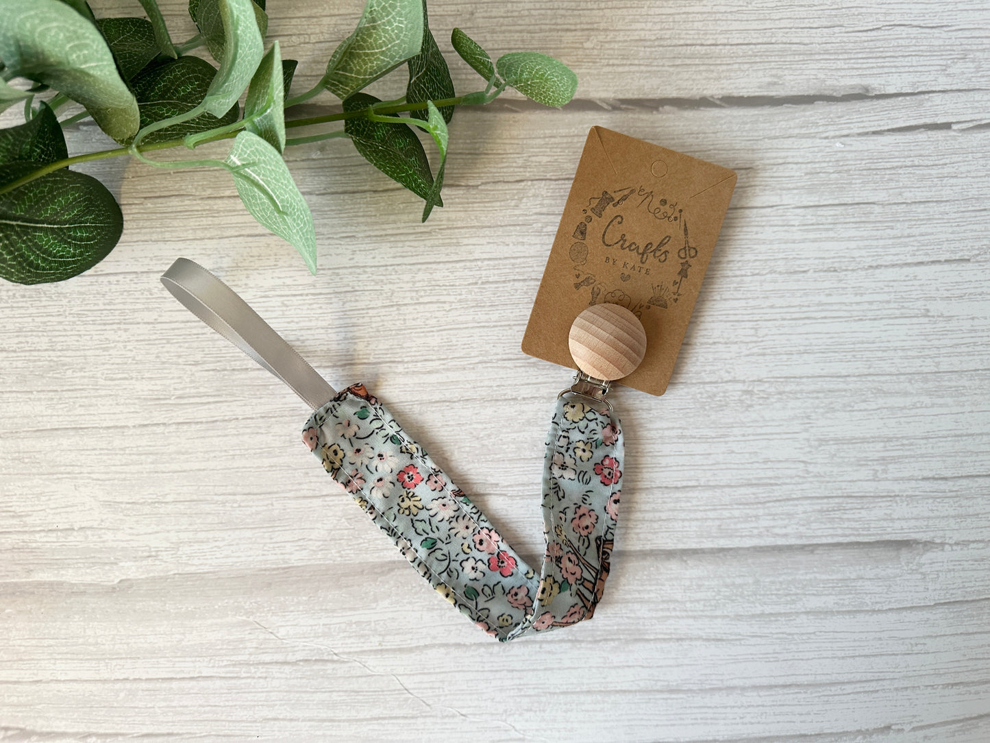 The "Cotton & Terry Towelling Baby Gift Set - Floral Bambi" by Crafts by Kate includes a floral cotton pacifier clip with a wooden button, elegantly attached to a "Messy Crafts" card. It sits on a white wooden surface adorned with fresh green leaves.