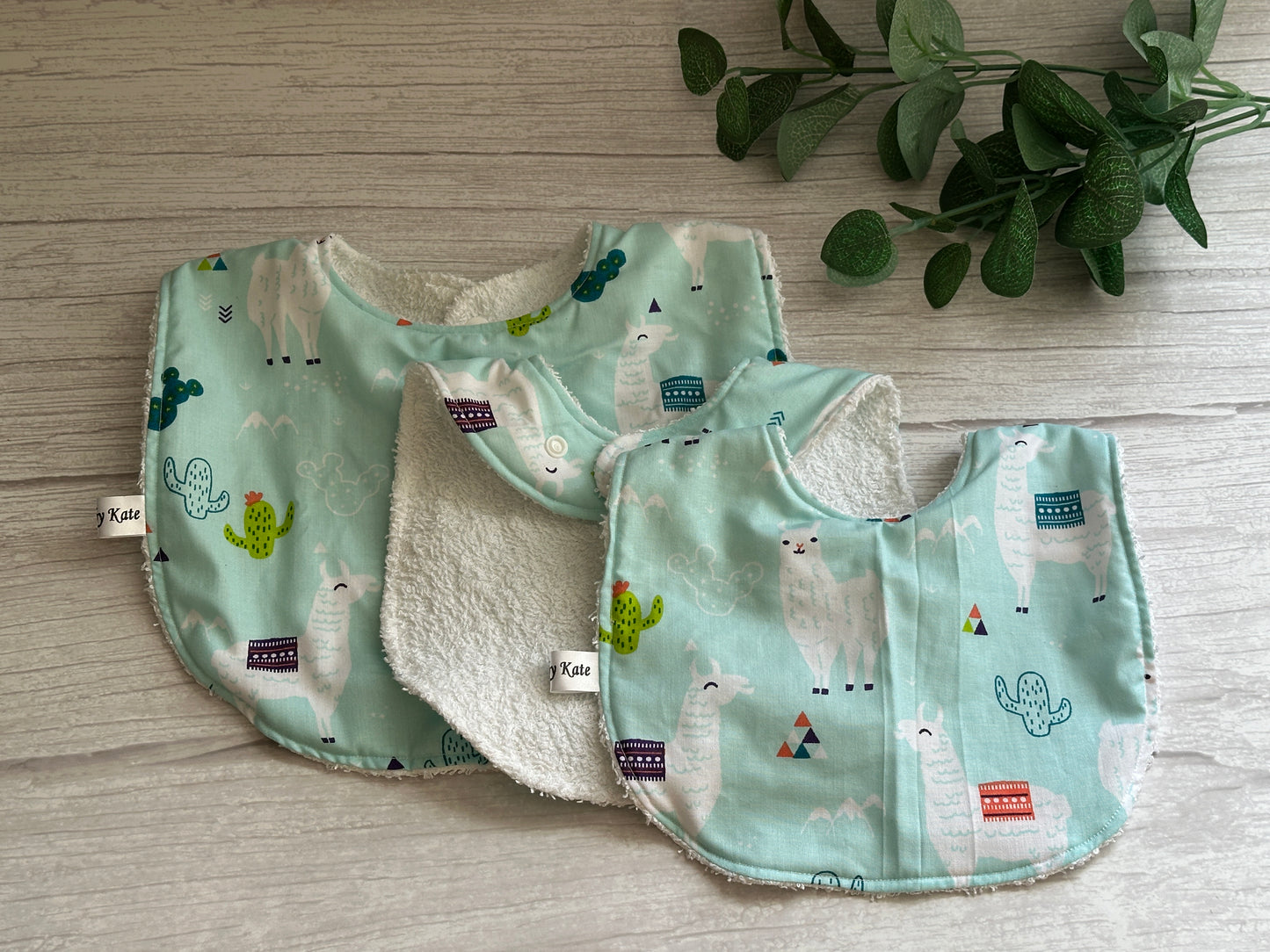 Two handcrafted "Llama Loves" baby bibs by Crafts by Kate, made of cotton and terry toweling, rest on a wooden surface. The charming scene is enhanced by a small leafy plant in the upper right corner, highlighting the softness and playful prints of these accessories.