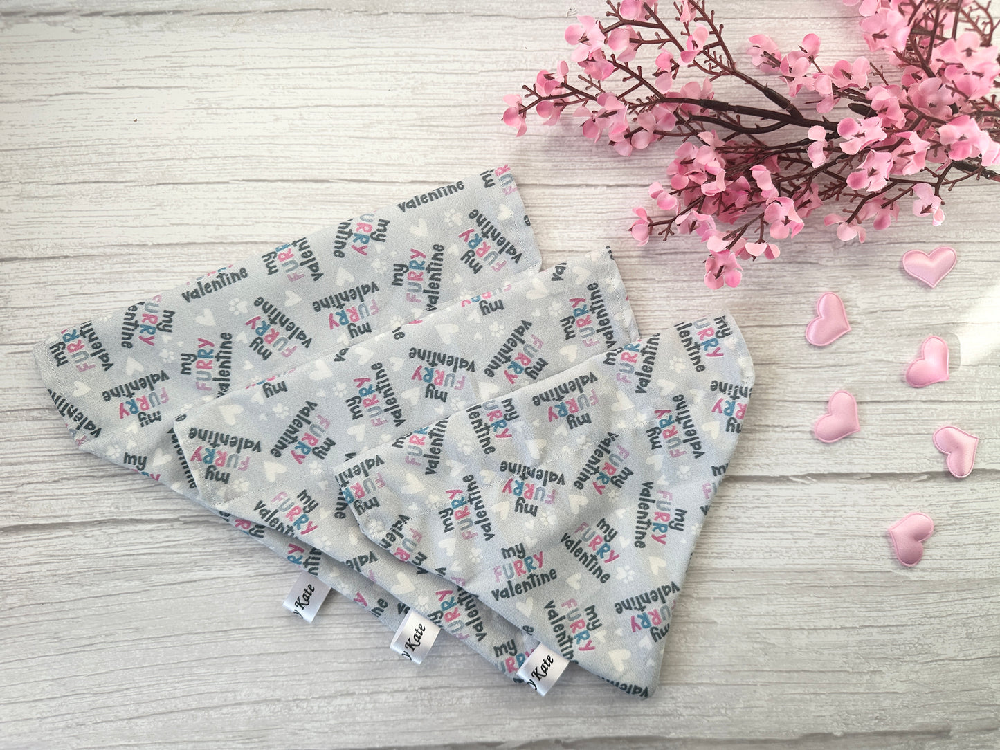 Four heart-shaped fabric coasters with "My Funny Valentine" patterns sit on a light wood surface. Nearby, pink flowers and heart-shaped decorations enhance the romantic theme, while the Cotton Pet Bandana - My Fluffy Valentine by Crafts by Kate adds charm with its soft texture.