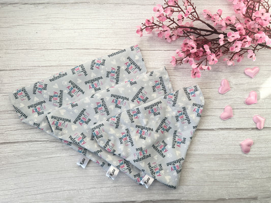 Four heart-shaped fabric coasters with "My Funny Valentine" patterns sit on a light wood surface. Nearby, pink flowers and heart-shaped decorations enhance the romantic theme, while the Cotton Pet Bandana - My Fluffy Valentine by Crafts by Kate adds charm with its soft texture.
