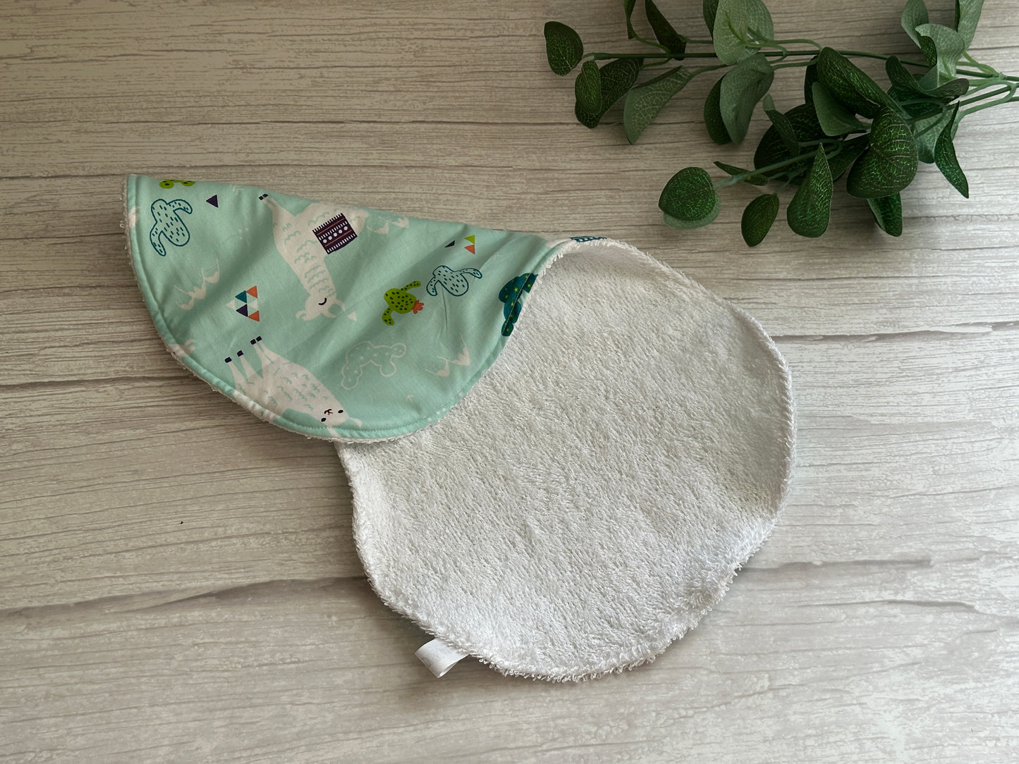 A charming, curved ultra-soft cotton bib from Crafts by Kate's "Llama Loves" collection rests on a wooden surface. One side showcases a colorful llama pattern while the reverse is white and textured. A small green plant adorns the corner, adding a touch of nature to this adorable accessory.