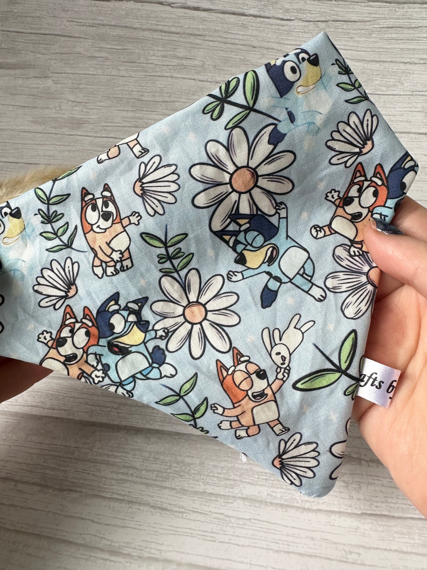 A hand displays the Cotton Pet Scrunchie Bandana - Bluey & Bingo from Crafts by Kate, showcasing a playful cartoon dog print surrounded by large white daisies and green leaves on a light blue fabric.