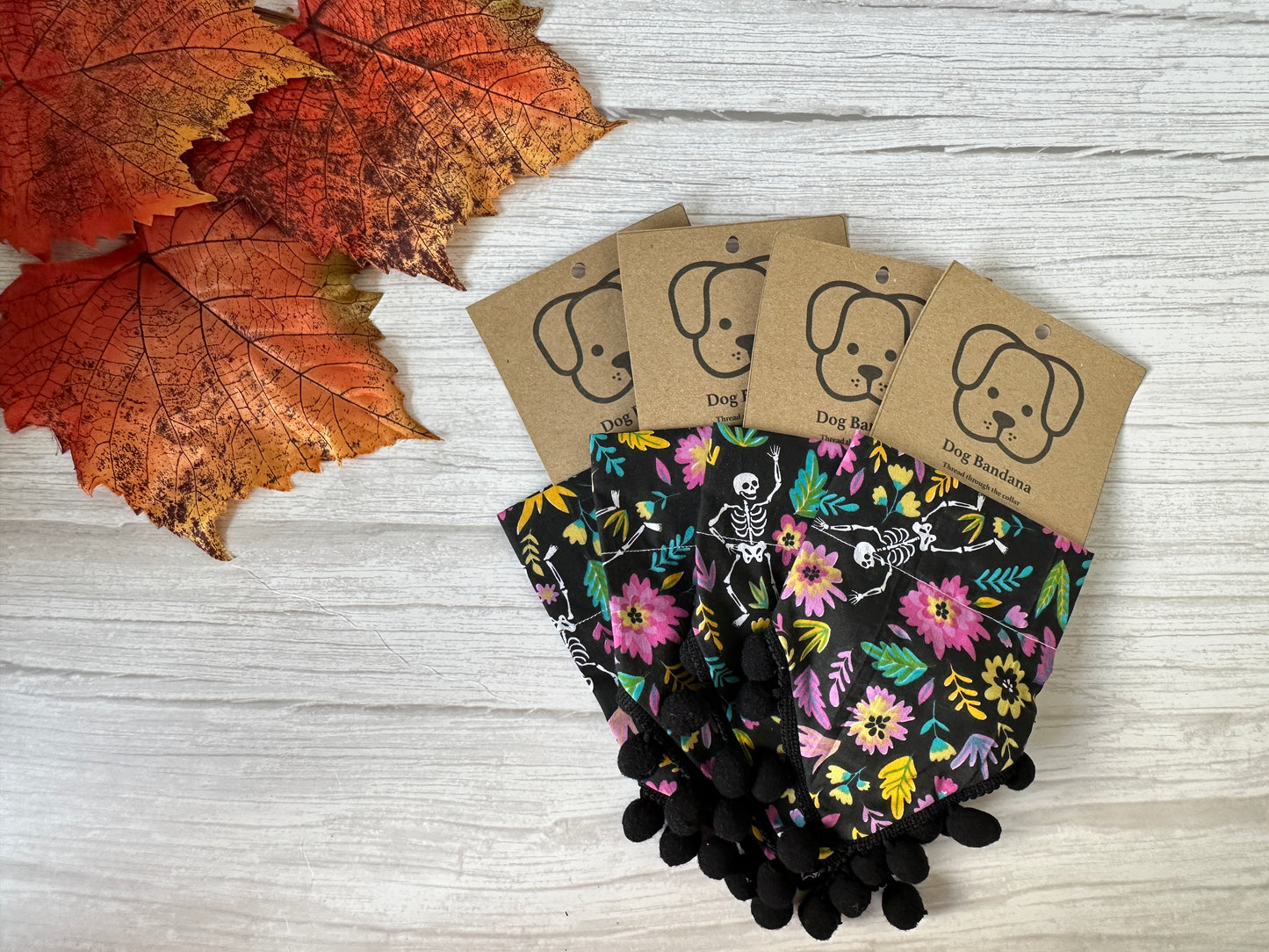 Four "Floral Skeletons with Pom Poms" cotton pet bandanas by Crafts by Kate are arranged on a white wooden surface with autumn leaves. Each features a collar attachment and a brown tag with a dog illustration and "Dog Bandana" text.