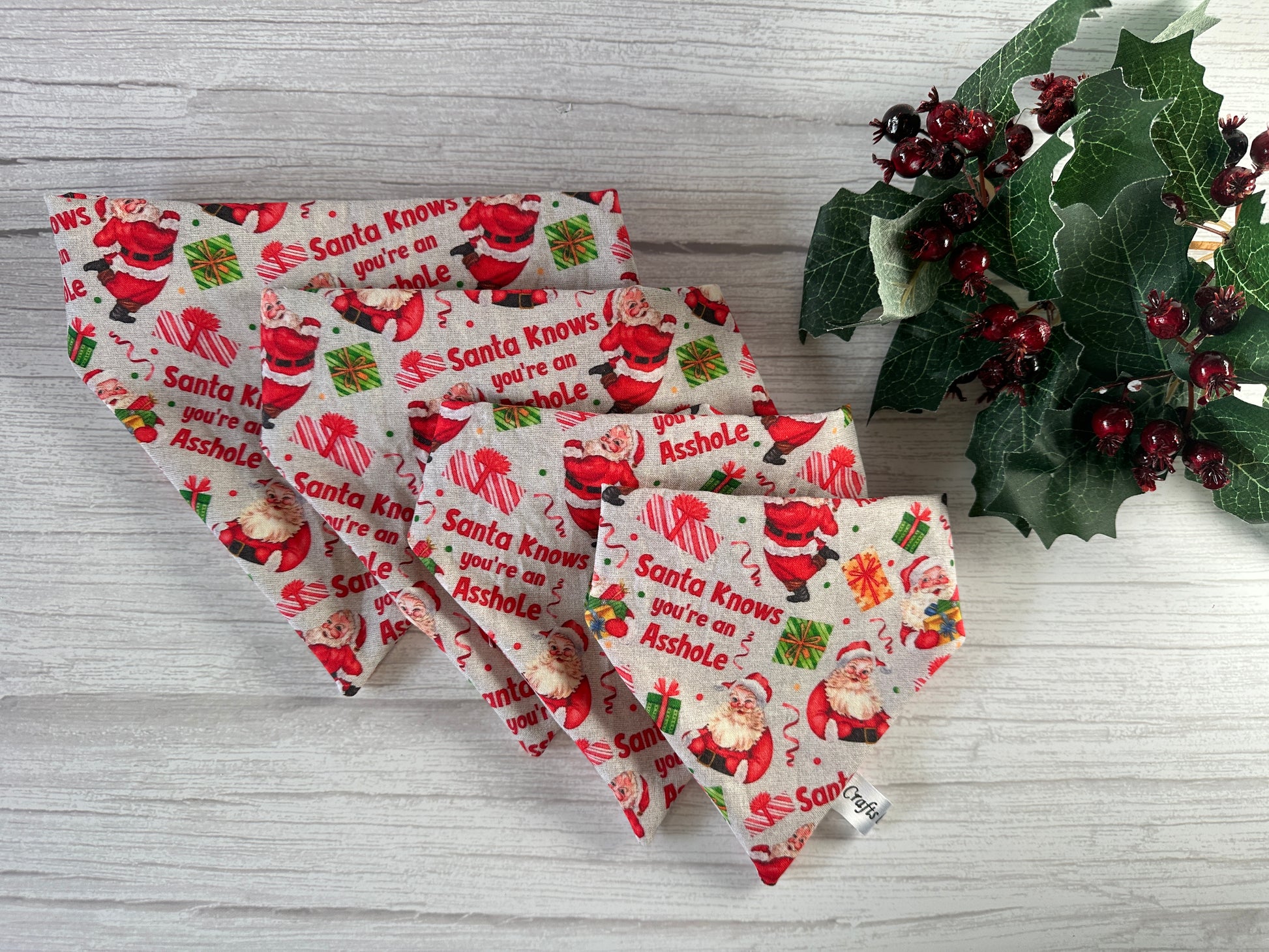 The Crafts by Kate Cotton Pet Scrunchie Bandana - Santa Knows, featuring “Santa knows you’re an asshole,” is displayed on a light wooden surface with holly and red berries decorating the side.