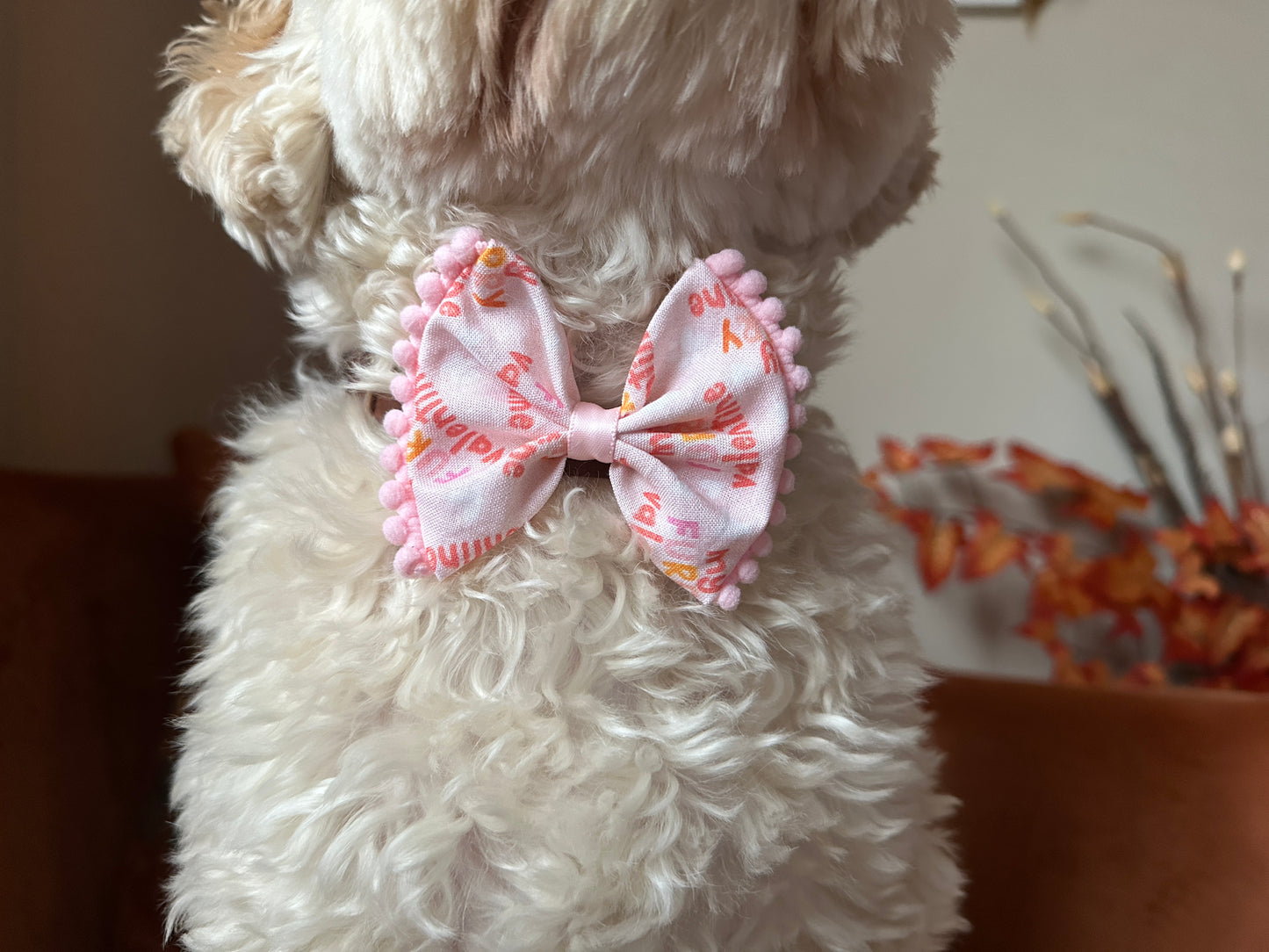 A fluffy dog is seen from behind, wearing the "My Fluffy Valentine" pink cotton pet bow by Crafts by Kate, featuring small pom-poms and visible text. The backdrop highlights warm autumn foliage.
