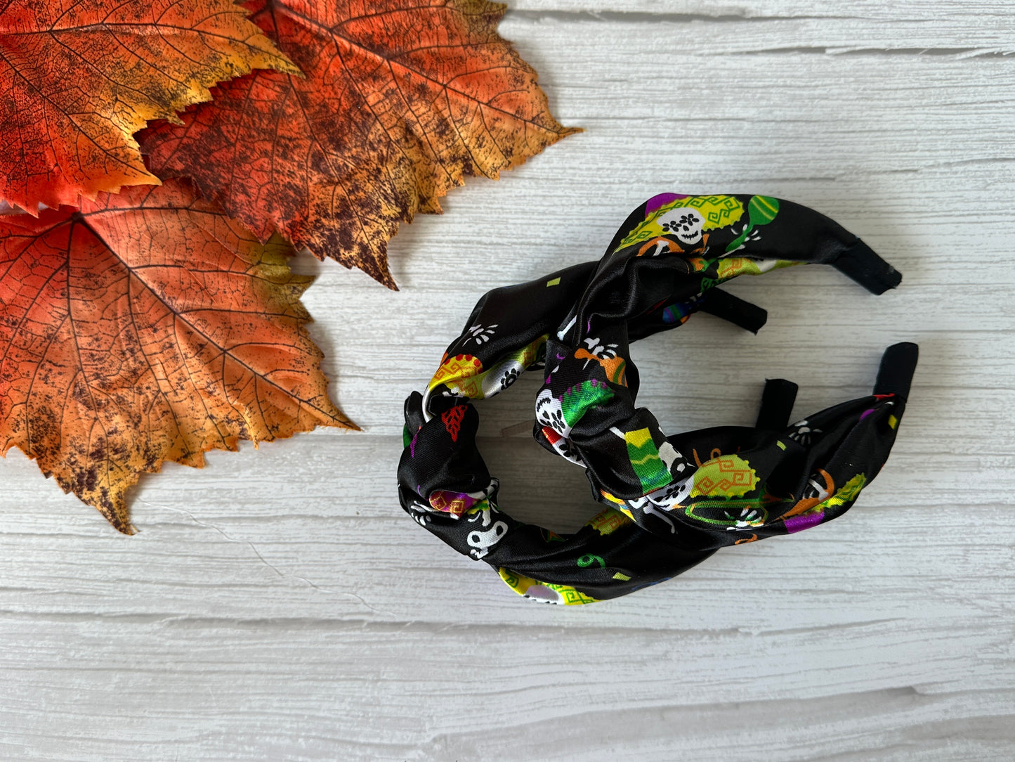 A pair of Crafts by Kate Silk Knotted Headbands – Day of the Dead, featuring colorful Halloween designs, rests on a white wooden surface beside orange autumn leaves.