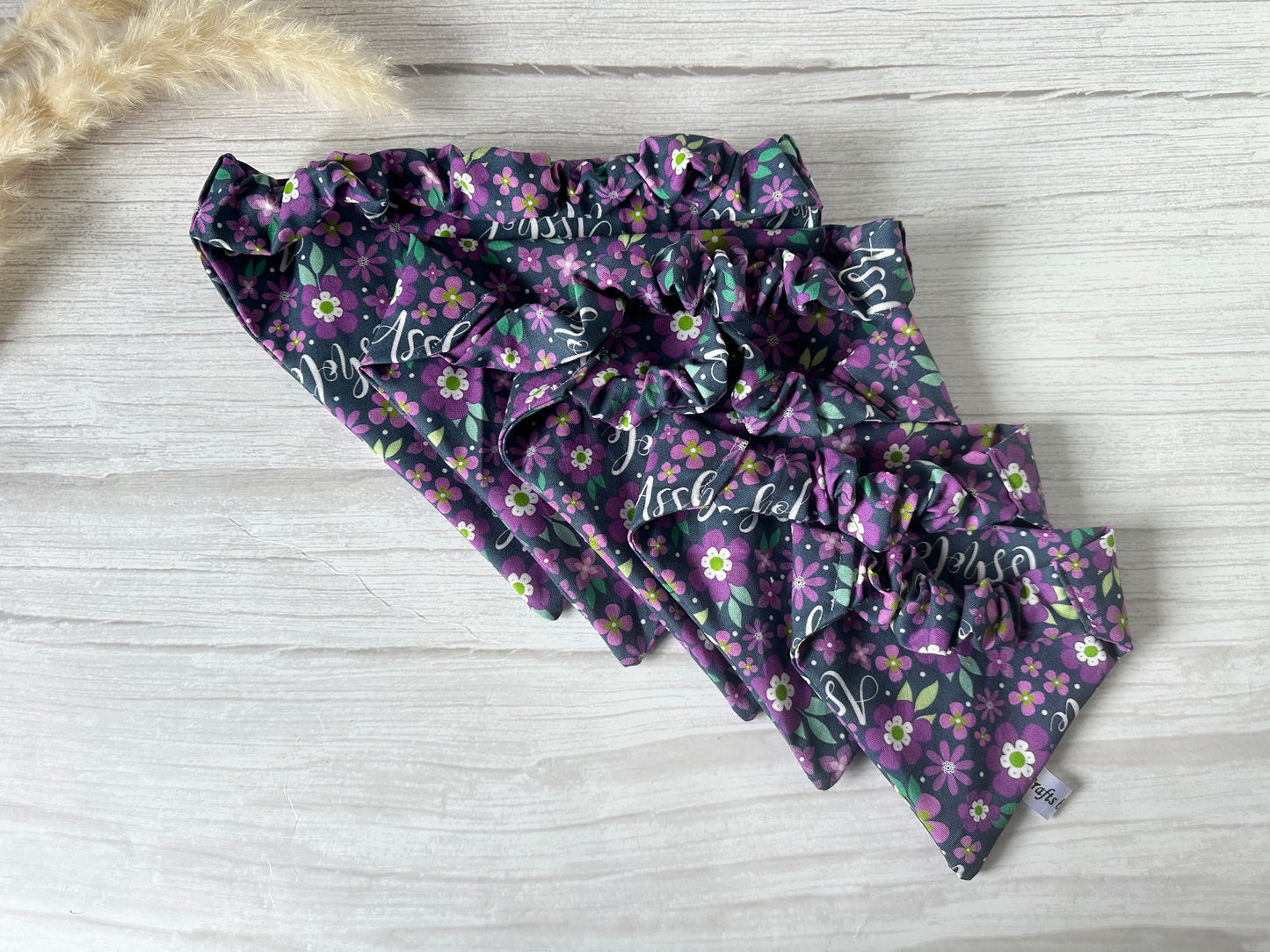 Crafts by Kate's Cotton Pet Scrunchie Bandana - Asshole features floral designs and script on dark purple. Displayed on a light wooden surface, various sizes are available. A small piece of dried grass in the top left corner adds a rustic touch.