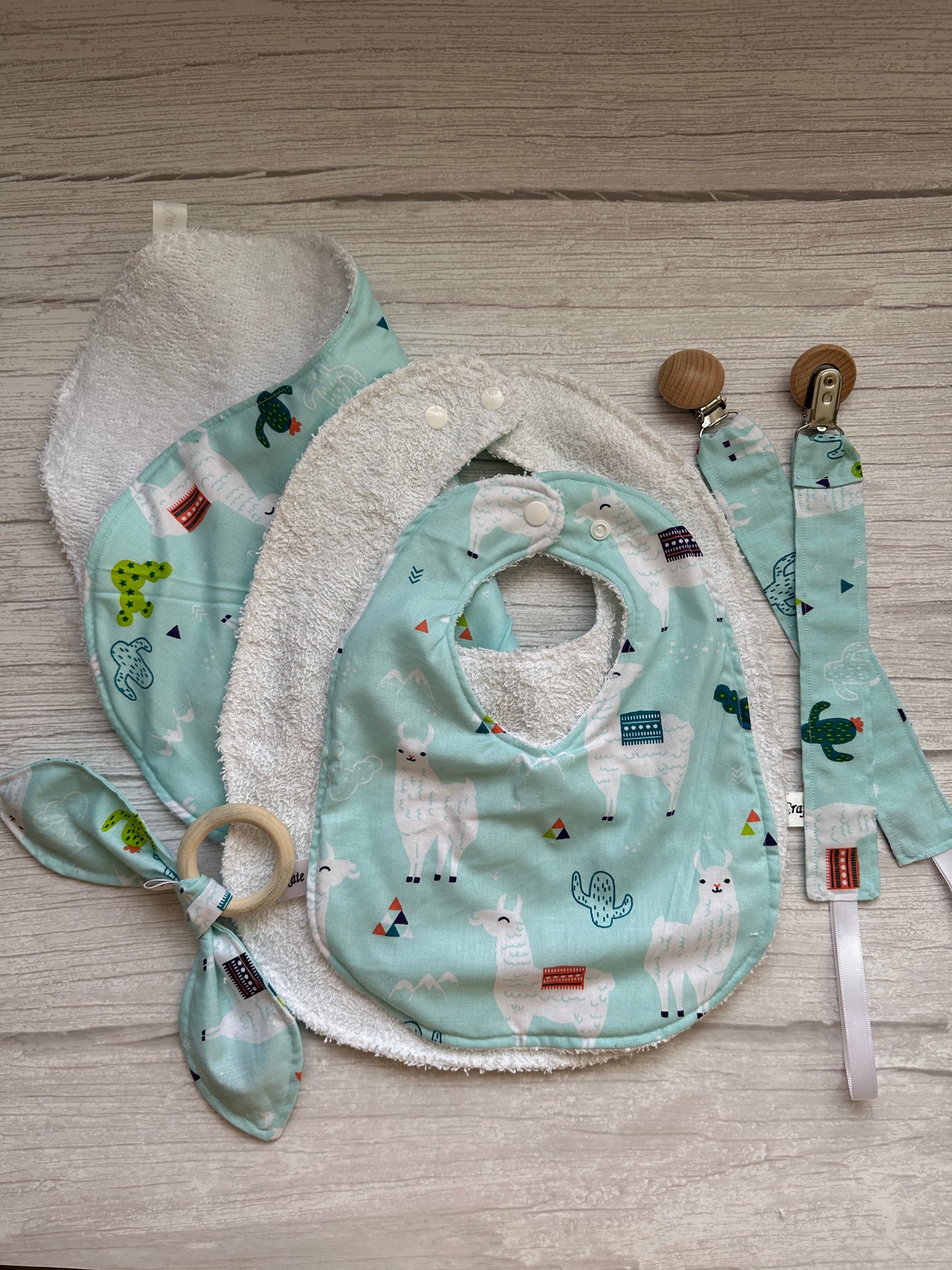 The "Llama Loves" set by Crafts by Kate showcases two ultra-soft cotton bibs with llama and cactus prints, a green pacifier clip, and a bamboo teething ring with a matching knotted cloth on a wooden background.