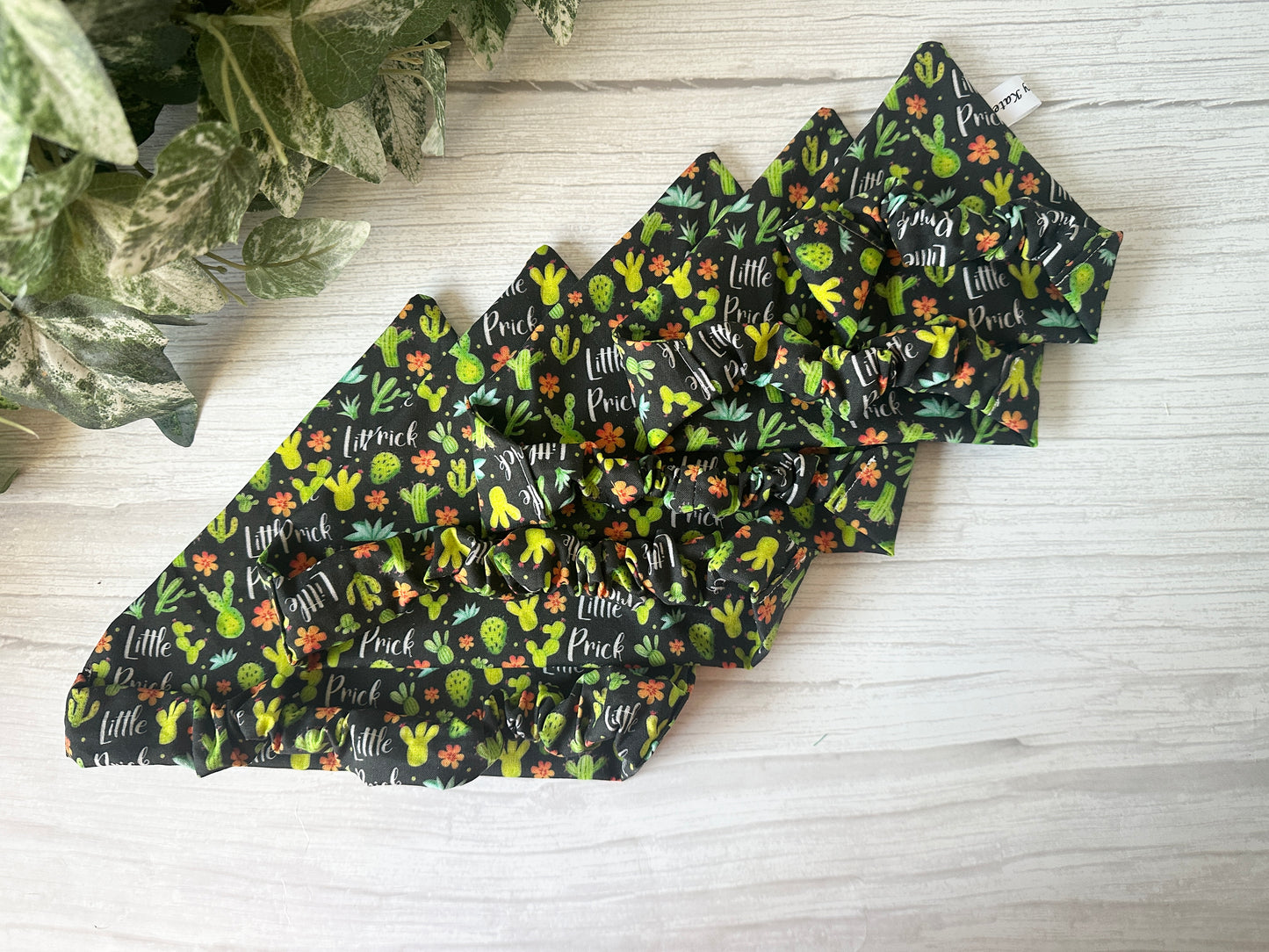 Five "Cotton Pet Scrunchie Bandana - Little Prick" accessories by Crafts by Kate, featuring cactus and floral prints, are fanned out on a light wood surface beside green leafy plants.