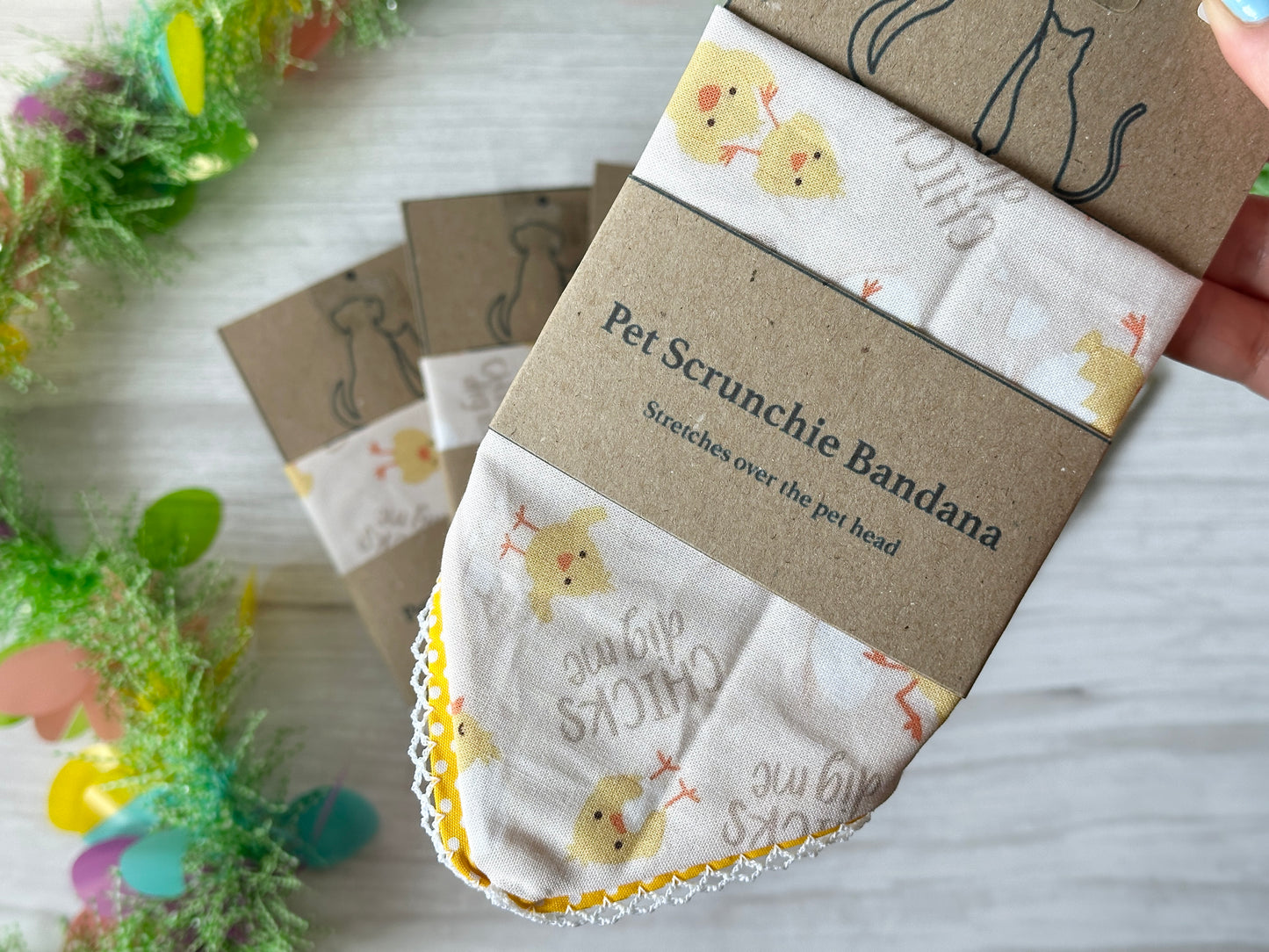 A hand presents the "Cotton Pet Scrunchie Bandana - Chicks Dig Me" from Crafts by Kate, made of handcrafted cotton with a playful chick design. In the background, more bandanas with colorful decorations are perfect for a special barkday celebration.