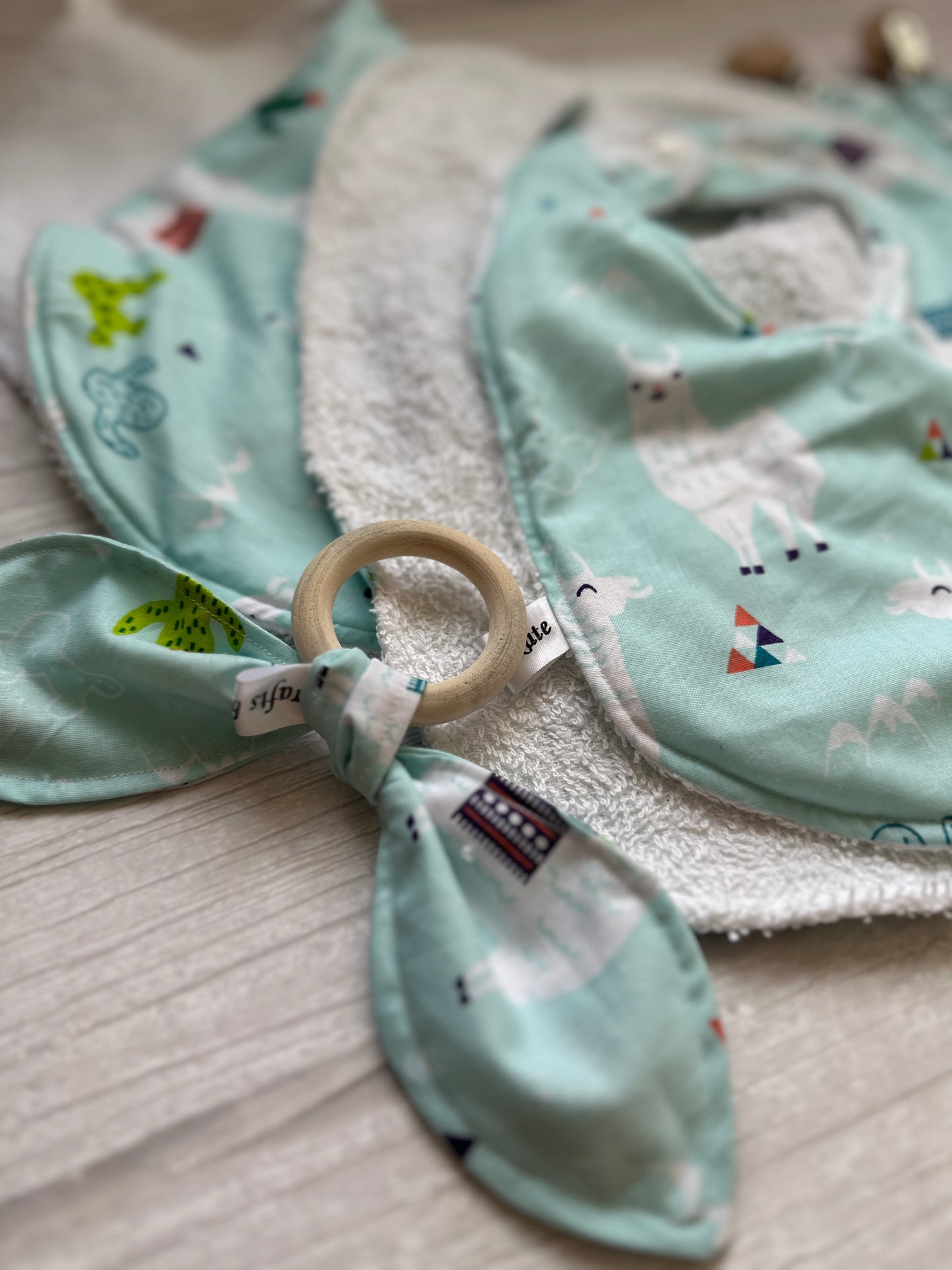 The "Llama Loves" baby set by Crafts by Kate features a soft blue cotton blanket with cartoon animals and a bamboo teething ring, displayed on a light wooden surface showcasing colorful llamas and tents.