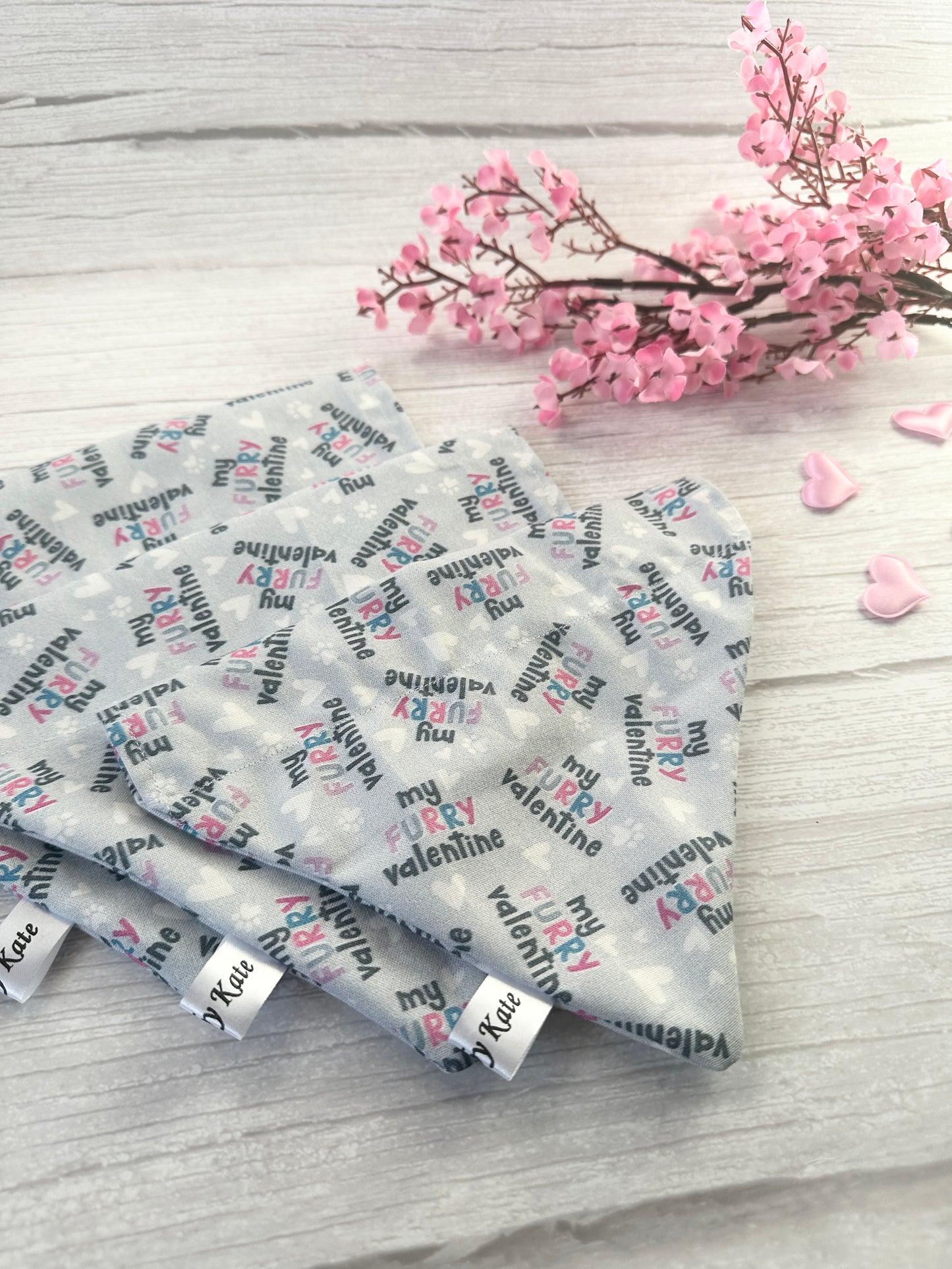 A light wood surface features gray square coasters with "My Crazy Valentine" in pink and black. The scene is romantically adorned with small pink hearts, cherry blossoms, and a Crafts by Kate Cotton Pet Bandana with the "My Fluffy Valentine" design.