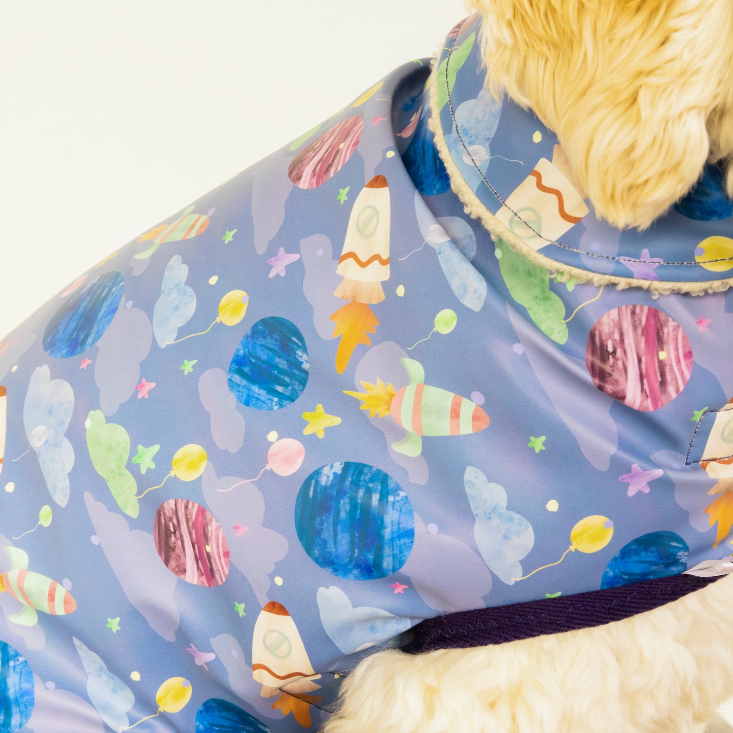A close-up of a light-colored dog wearing the Crafts by Kate Space Adventure Pet Raincoat, featuring a waterproof design and colorful planets, stars, clouds, and rockets on a lavender background.