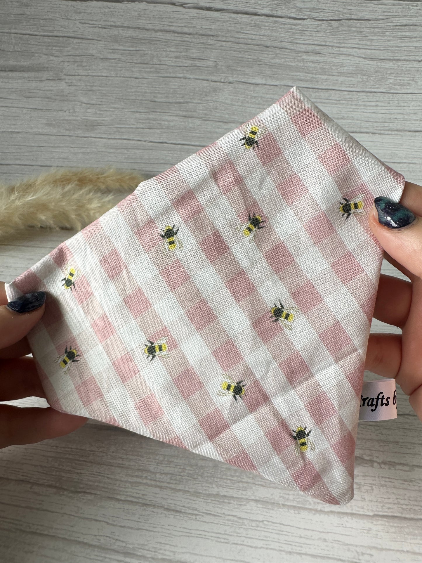 Hands holding a Cotton Pet Scrunchie Bandana - Pink Gingham Bees by Crafts by Kate, showcasing its charming bee patterns on a pink gingham fabric against a light wooden background.