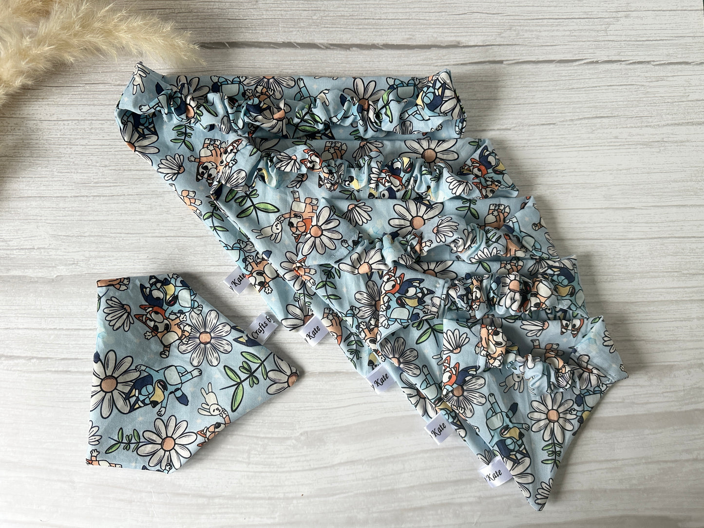 The Crafts by Kate Cotton Pet Scrunchie Bandana - Bluey & Bingo set, featuring a light blue design with white flowers, orange accents, and green leaves, includes five coordinating scrunchies. Displayed on a white wooden surface, it creates a delightful spring arrangement.