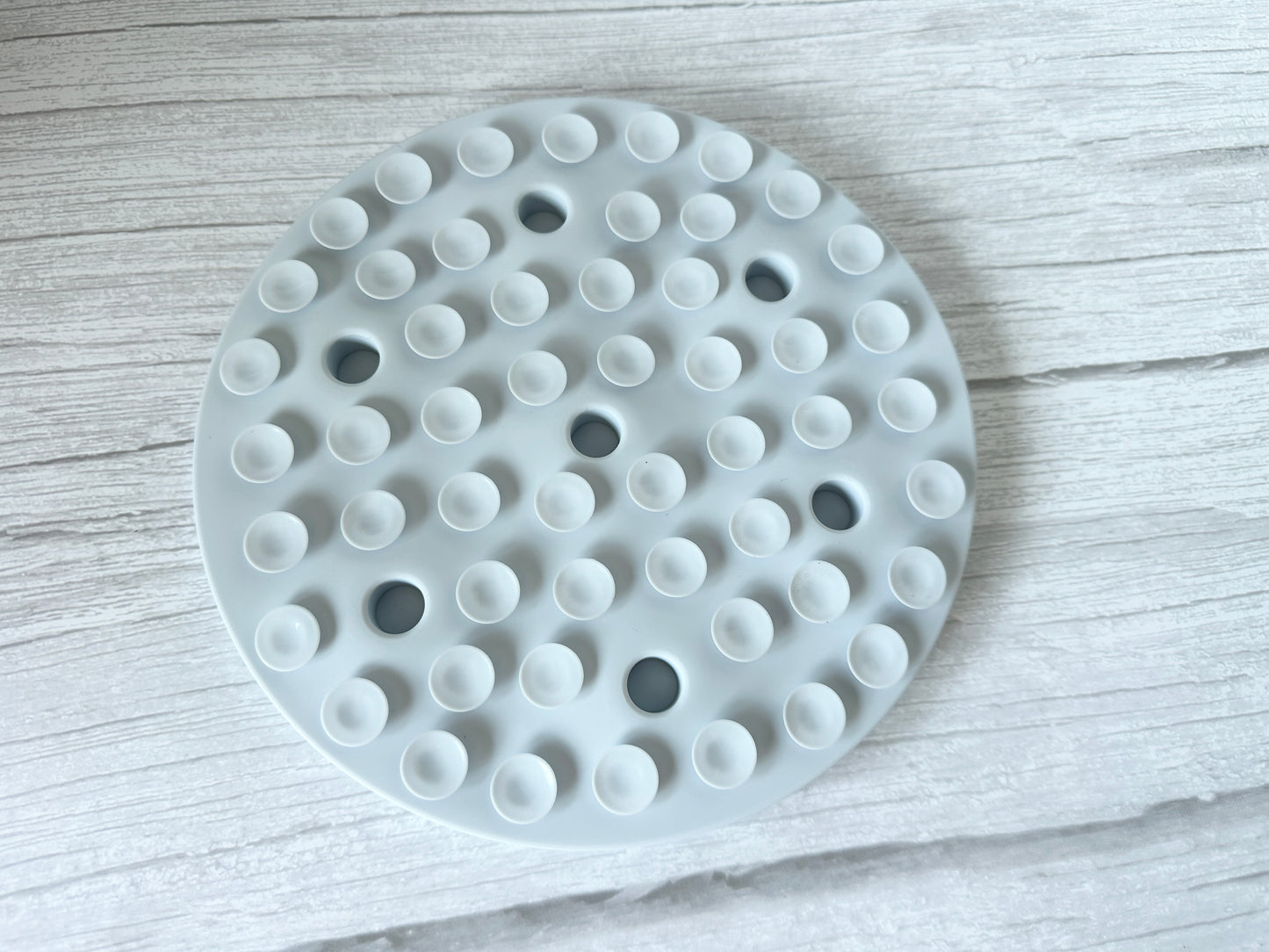 The Slow Feeder Pet Mat by Crafts by Kate is a round, white silicone mat with evenly spaced suction cups, set on a textured, light-colored wood surface. Its minimalist design features multiple small bumps that aid pet digestion and ensure easy cleaning.