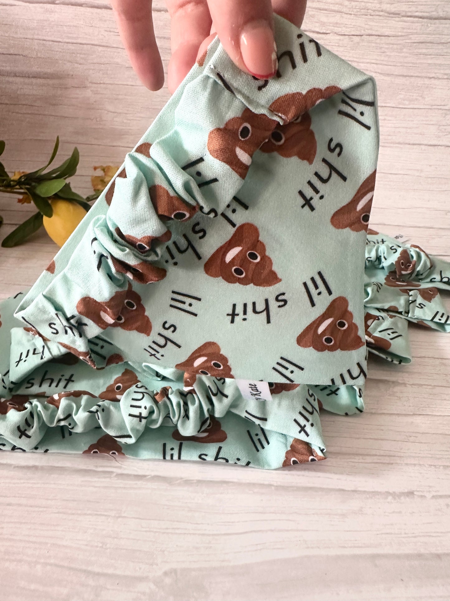 A hand holds up the "Lil Shit" Cotton Pet Scrunchie Bandana by Crafts by Kate, featuring cartoon poop emojis and text, beside a matching scrunchie. A lemon and greenery decorate the background.
