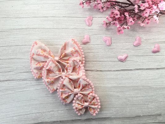 The "My Fluffy Valentine" cotton pet bow by Crafts by Kate, with its red "Happy Valentine's Day" text, sits elegantly on a light wood surface. The scene is charmingly completed with pink cherry blossom branches and tiny heart-shaped decorations.
