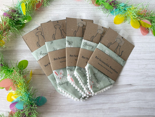 Crafts by Kate's "Snuggle Bunny" Cotton Pet Scrunchie Bandanas, made with pastel green fabric and pom-pom trim, are neatly arranged on a wooden surface, surrounded by colorful sprigs and Easter eggs, emphasizing the "Chicks Dig Me" charm of this cute ensemble.