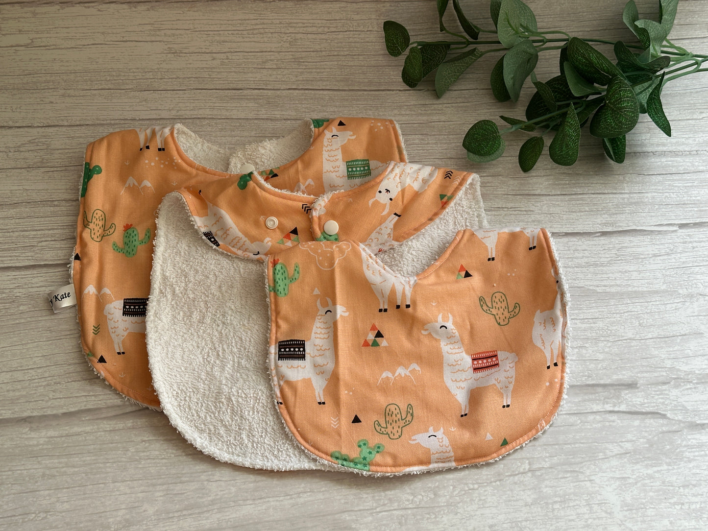 Three orange Cotton & Terry Towelling Baby Sets - Llama Loves by Crafts by Kate, featuring ultra-soft llama and cactus patterns, are stacked on a wooden surface. Made from handcrafted cotton for gentle comfort, with green leaves partially visible in the top right corner.