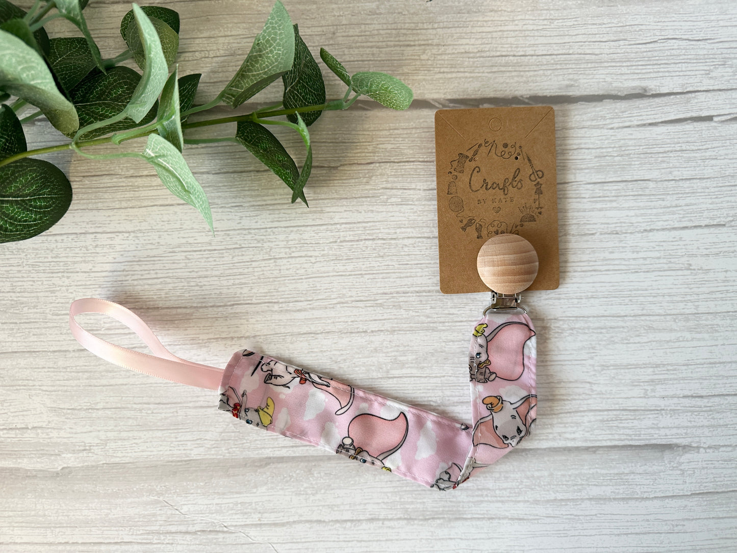 The Cotton & Terry Towelling Baby Gift Set - Dumbo in pink by Crafts by Kate features a pink pacifier clip with a cartoon elephant, attached to a wooden clip. Its charm is enhanced by green foliage and a decorative cardboard tag, highlighting this handcrafted ultra-soft cotton item.