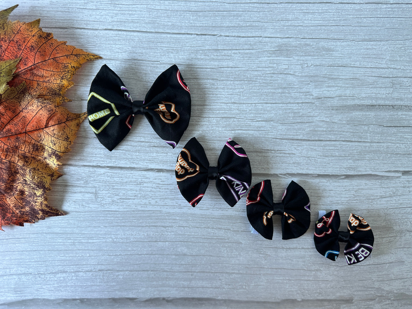 Four Crafts by Kate Cotton Pet Bows with collar attachments, each larger than the last and featuring glow-in-the-dark Signs designs, are arranged on a light gray wooden surface with a colorful autumn leaf to the left.