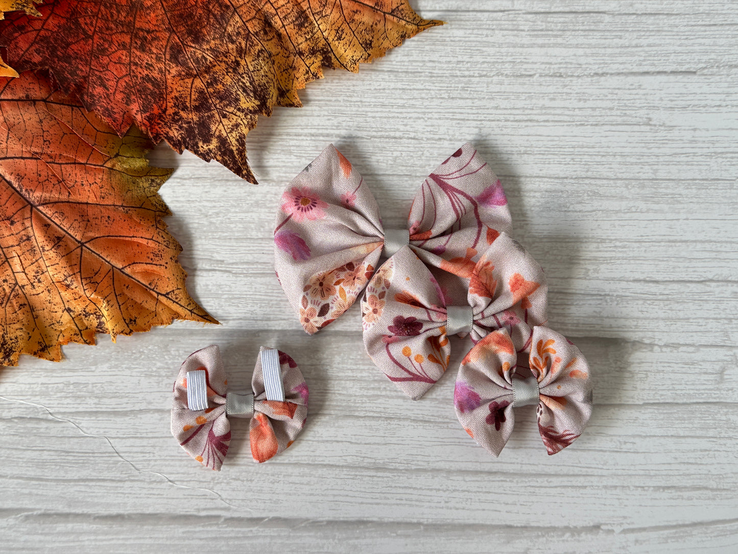 The Cotton Pet Bow with collar attachment - Woodland Floral by Crafts by Kate features pink, orange, and purple floral patterns in various sizes, displayed on a white wooden surface beside autumn-colored artificial leaves.