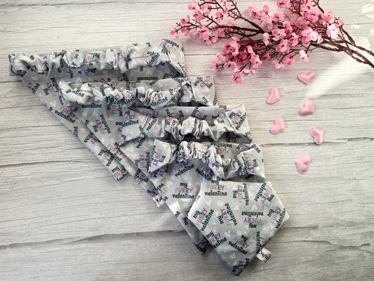 On a light wooden surface, four gray "My Furry Valentine" scrunchie bandanas from Crafts by Kate's "Cotton Pet Scrunchie Bandana - My Fluffy Valentine" collection lay alongside pink blossoms and decorative hearts, adding a romantic touch.