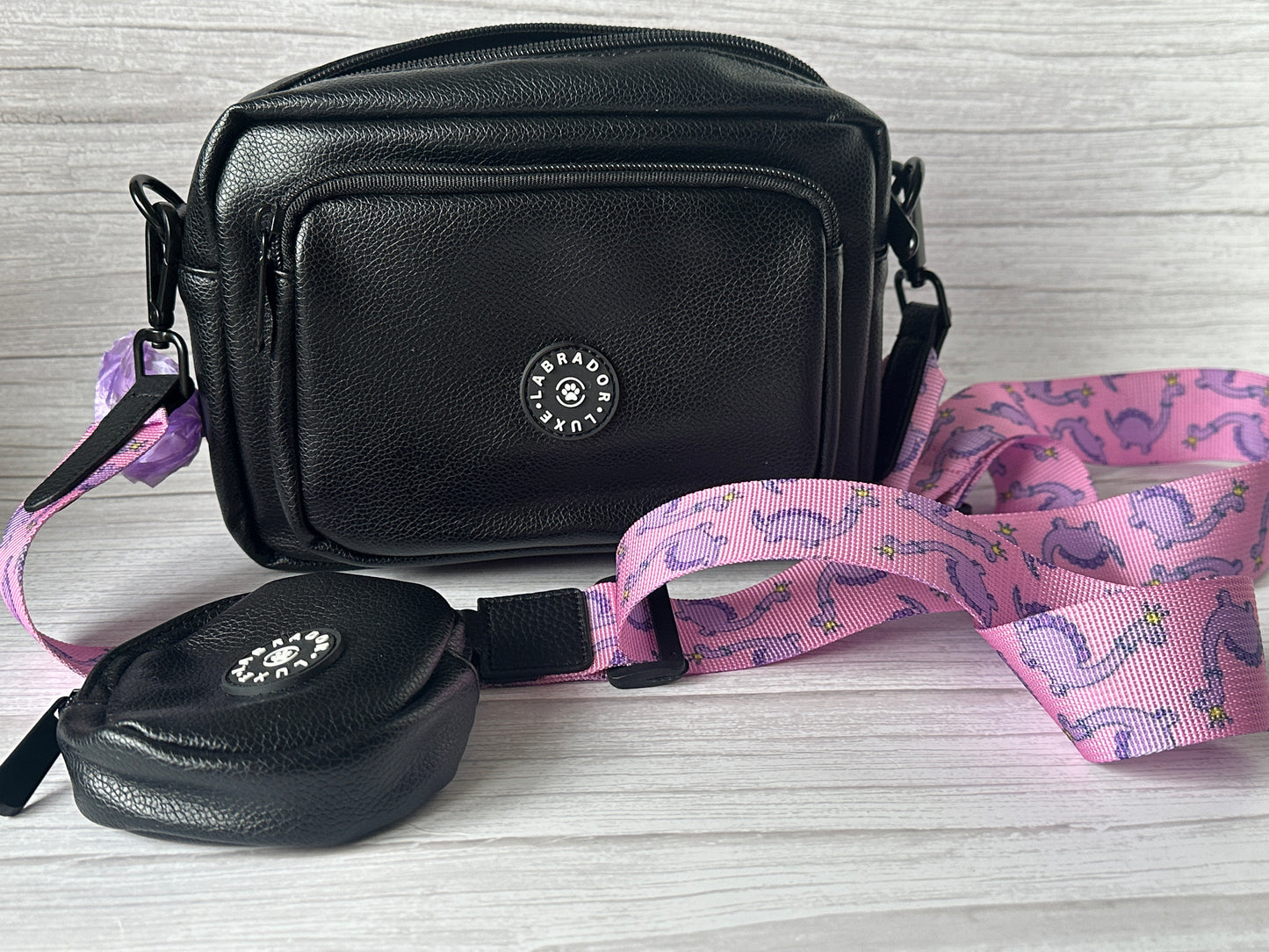 The Millie Dog Walking Bag by Crafts by Kate is a black vegan leather bag with a small matching treat pouch and a detachable pink strap featuring purple dinosaurs, shown on a light wooden surface.