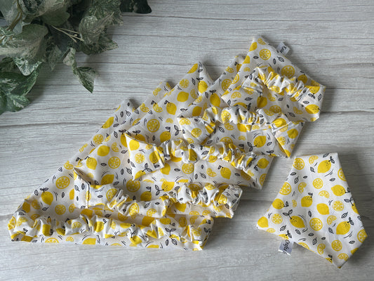 Four yellow and white "Lemons lovers" cotton accessories by Crafts by Kate—including three ruffled bandanas in different sizes and a stretchable pet scrunchie—are displayed on light wood beside green artificial leaves.