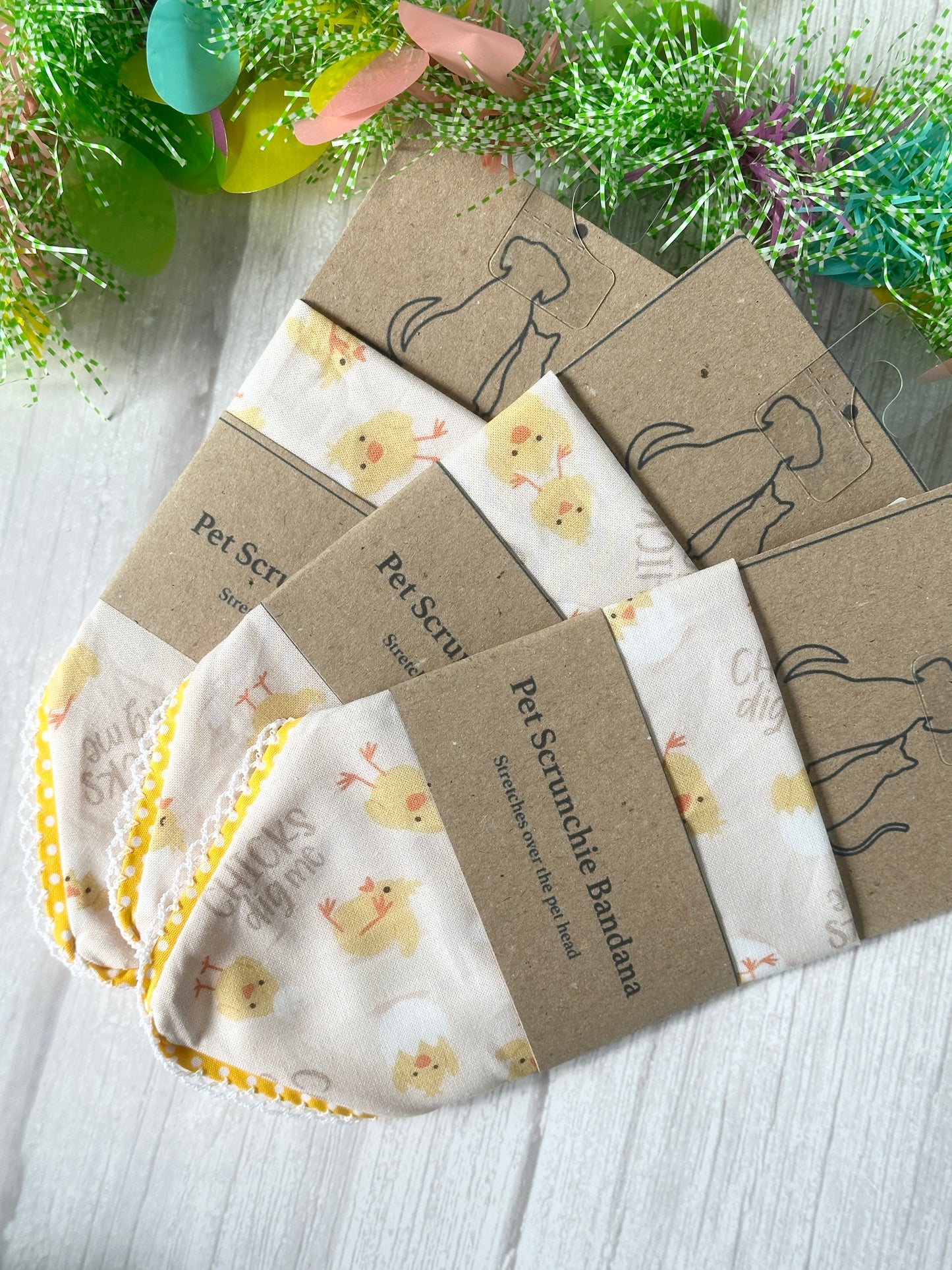 Three handcrafted Cotton Pet Scrunchie Bandanas—Chicks Dig Me design by Crafts by Kate—are wrapped in brown paper labels and set on a light wooden surface, with colorful decorative elements adorning the top and bottom.