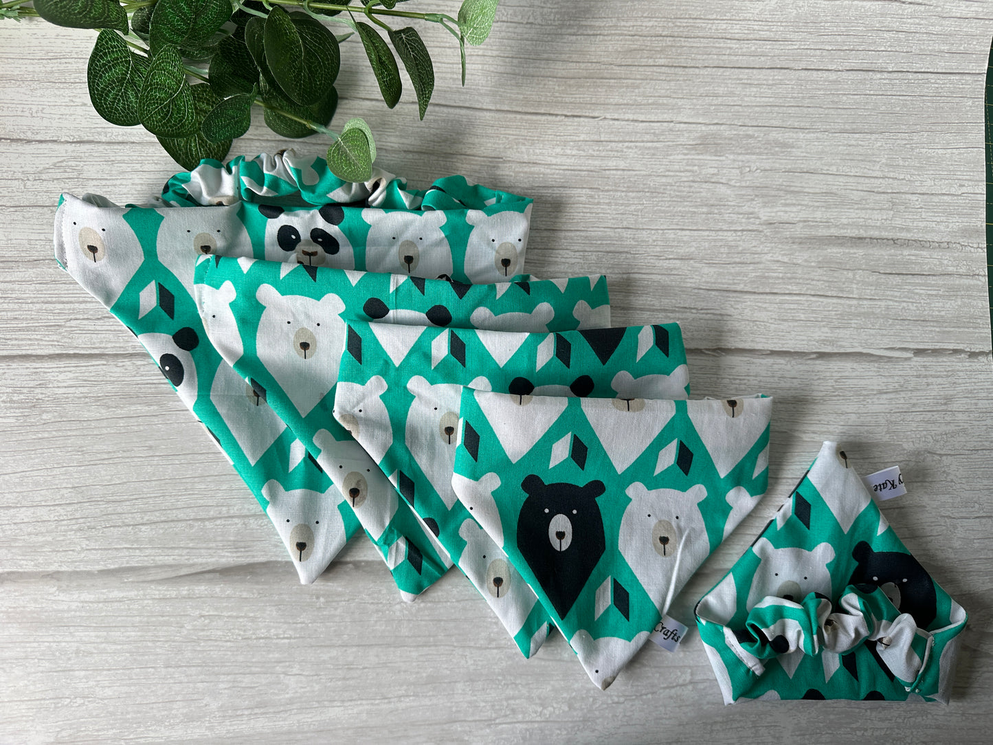 Five green and white patterned "It’s My Barkday" bandanas by Crafts by Kate, featuring bear designs, arranged fan-shaped from small to large next to matching pet scrunchies. A leafy plant peeks in the upper left on a wooden background, ideal for Bear Lovers Fabric enthusiasts.