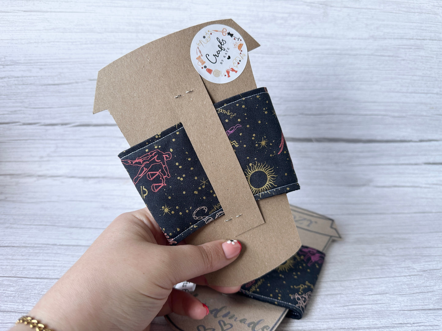A hand holds the "Cotton Reusable Cup Cozy - Horoscope Sky" by Crafts by Kate, featuring astral designs of stars, moons, and constellations. The eco-friendly cozy is on a brown card with a round sticker on a light wood surface.