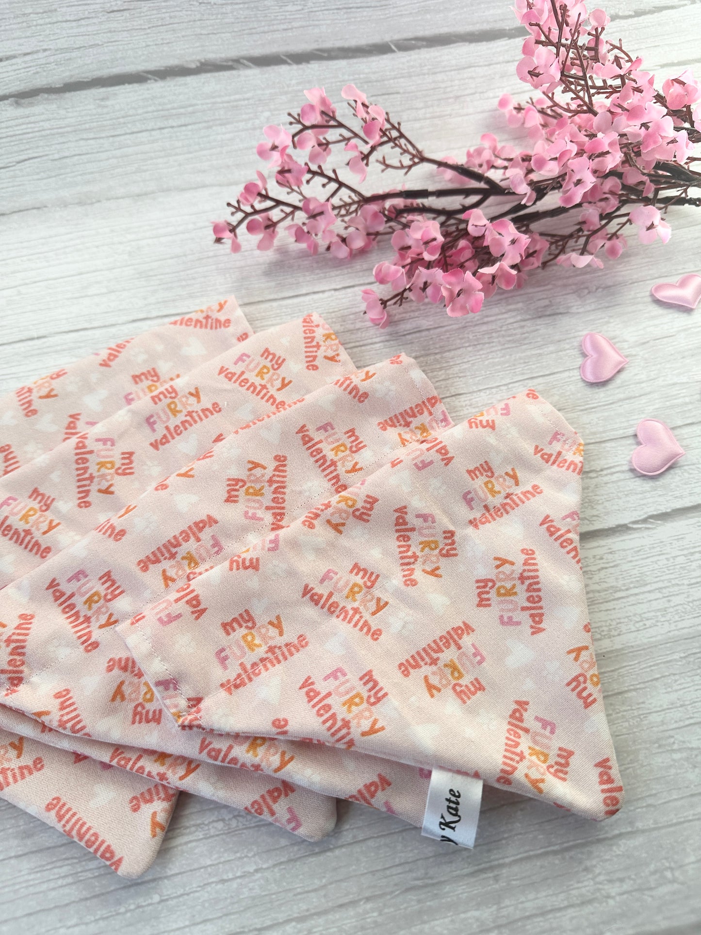 The Cotton Pet Bandana by Crafts by Kate, named "My Fluffy Valentine," features a charming design with red and orange text on a light wooden surface. Pink flowers and heart-shaped decorations in the top right corner add a delightful touch.
