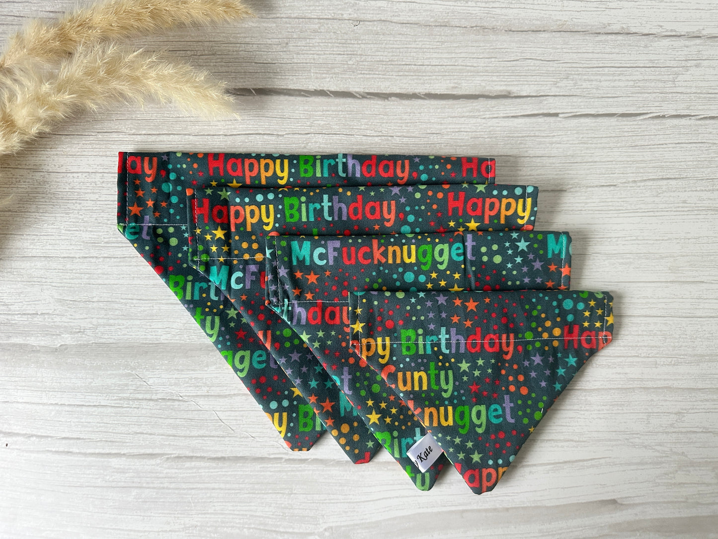 Crafts by Kate’s Cotton Pet Bandana, featuring the "Happy Birthday C Mcnugget" design, showcases playful text on colorful fabric. With a collar attachment and rustic wooden backdrop enhancing its vibrancy, it is perfect for giving your pet a festive flair.
