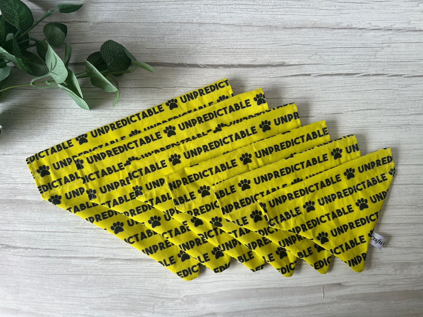 Seven Crafts by Kate "Unpredictable" Cotton Pet Bandanas with collar attachment, featuring black text and paw prints, are fanned out on a light wood surface with green leaves in the corner.