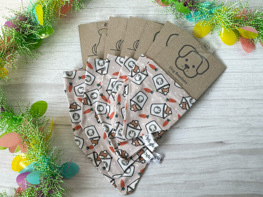 Five handcrafted "Easter Pupcup" cotton pet bandanas by Crafts by Kate, featuring cartoon designs and tags with dog illustrations, are fanned out on a light wood surface. Vibrant decorative foliage and Easter Pupcup Fabric eggs are artfully arranged around these charming accessories.