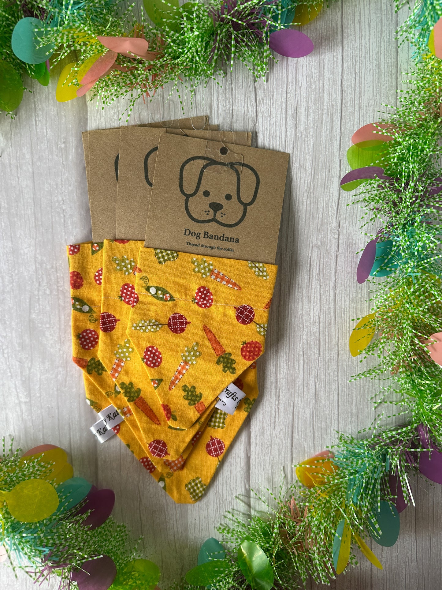 Crafts by Kate features three vibrant Cotton Pet Bandanas with collar attachments, adorned with vegetable patterns on a light wood surface. They're framed by a colorful garland and come packaged with an adorable dog illustration.