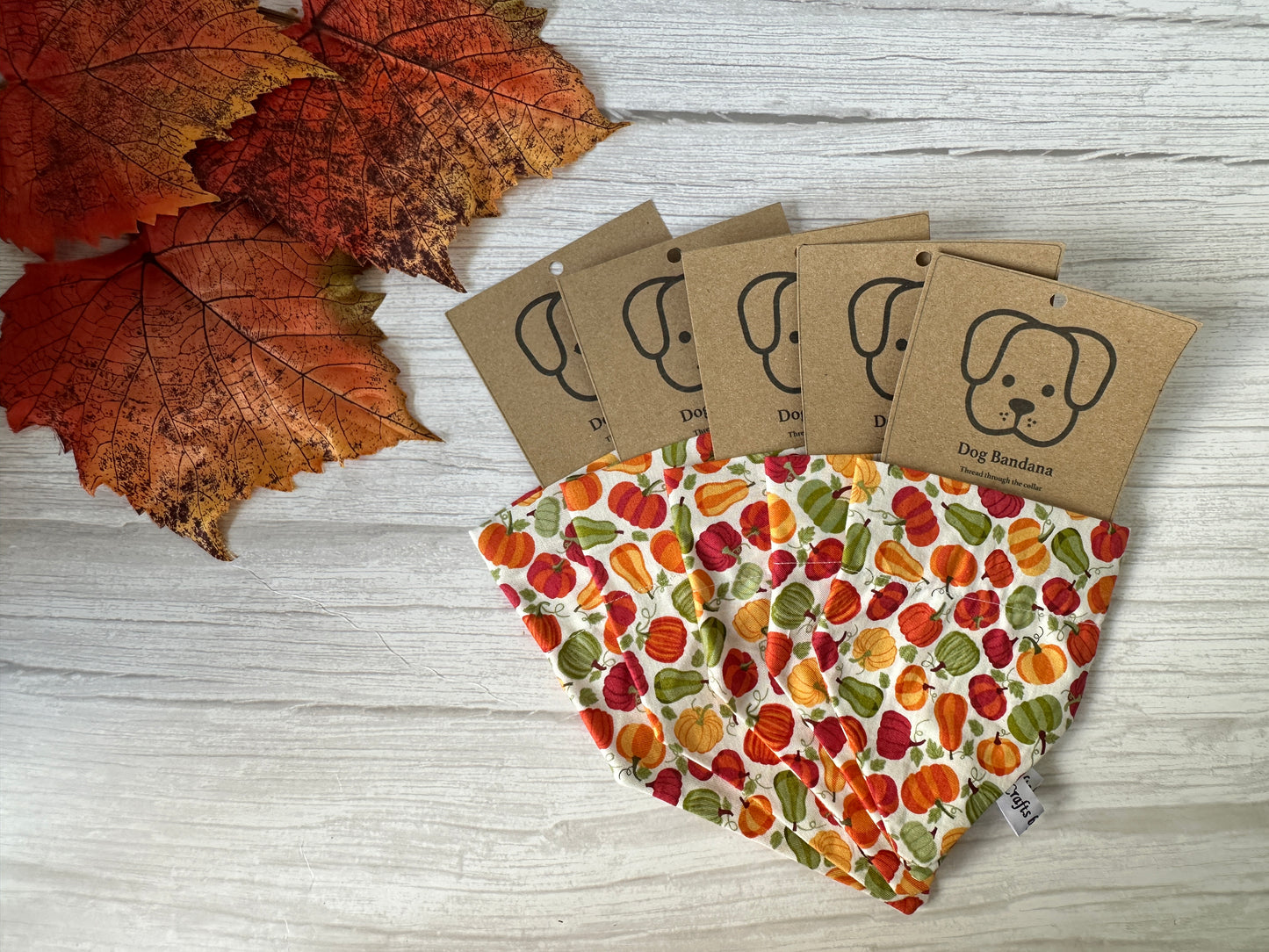 Four Crafts by Kate "Cotton Pet Bandana with collar attachment - Pumpkin Love" bandanas are displayed on light wood near autumn leaves. Each features a brown tag with a simple dog face and the words "Dog Bandana.