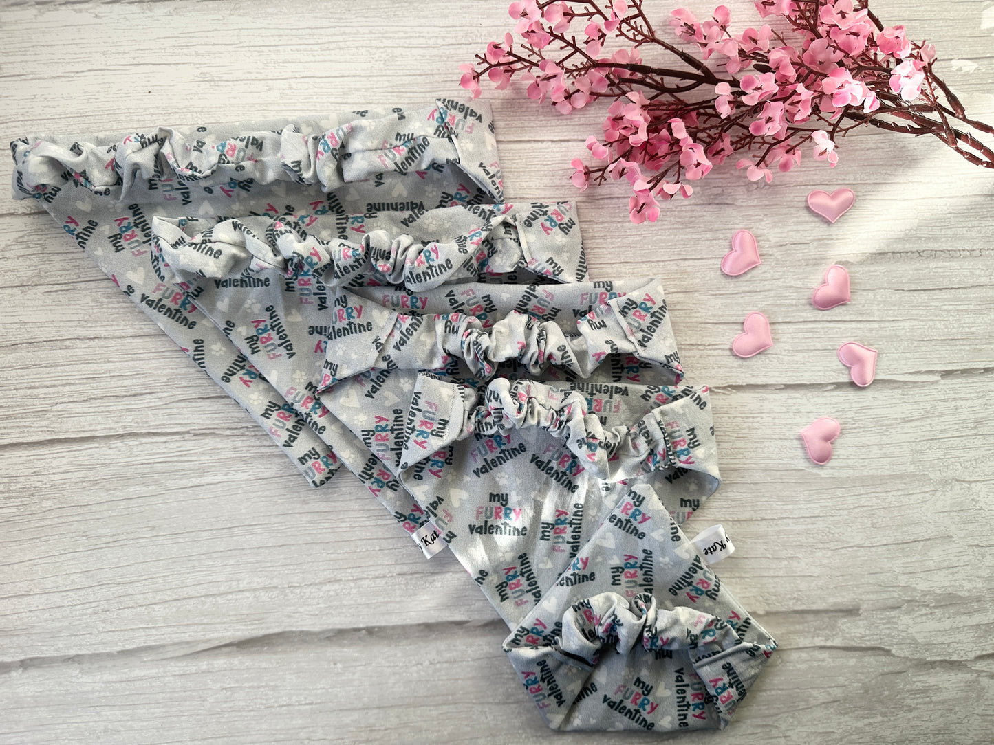 Four gray "My Furry Valentine" bandanas are fanned out on wood, accompanied by pink cherry blossoms, heart-shaped decorations, and a handcrafted Crafts by Kate cotton pet scrunchie named "My Fluffy Valentine.