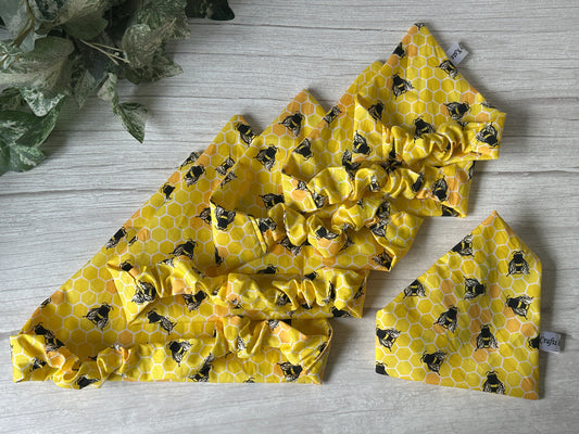 The Cotton Pet Scrunchie Bandana - Beehive Haven by Crafts by Kate features a cheerful yellow honeycomb pattern with black bees, displayed on a light wood surface with green foliage—charming pet accessories for any occasion.