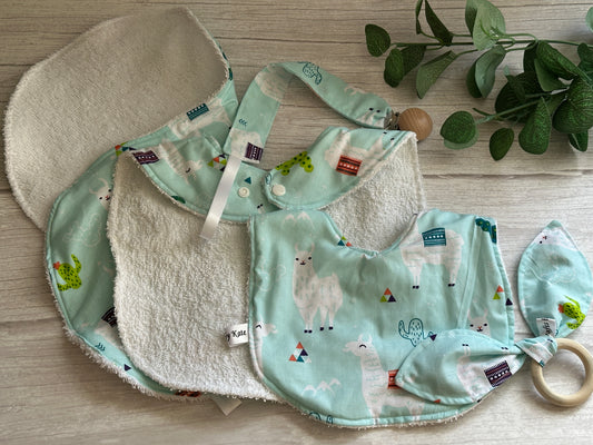 The marvelous Crafts by Kate "Cotton & Terry Towelling Baby Sets - Llama Loves" rests on a light wooden surface, showcasing handcrafted llama print cotton bibs and an ultra-soft pacifier clip. A leafy plant in the corner ties together this charming pastel-toned ensemble.