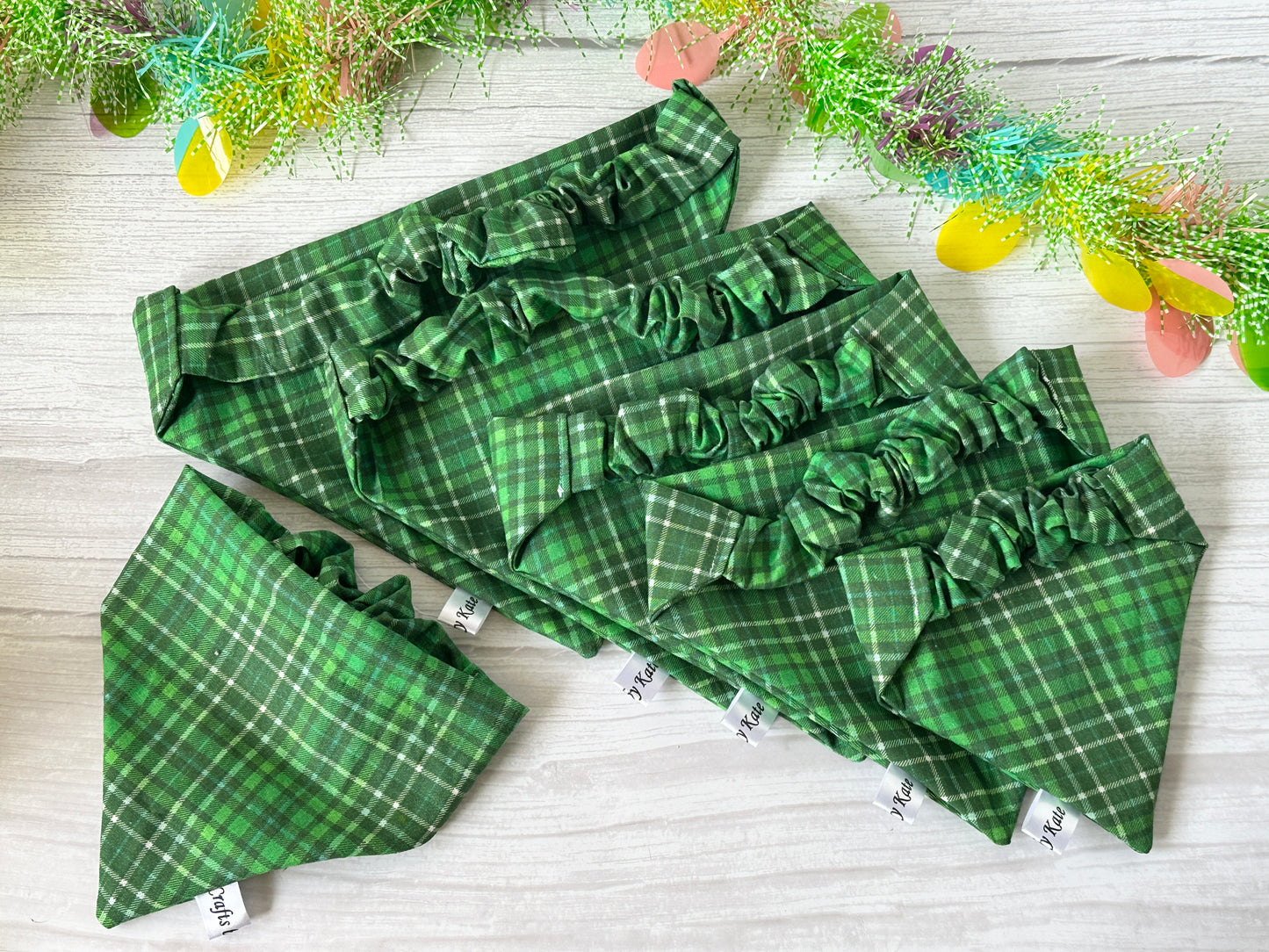 The Cotton Pet Scrunchie Bandanas - Bluey & Bingo by Crafts by Kate are displayed on a light wooden surface. Five bandanas form a cascading pattern, complemented by decorative greenery and colorful paper eggs in the background, showcasing their ruffled edges and spring tartan fabric.