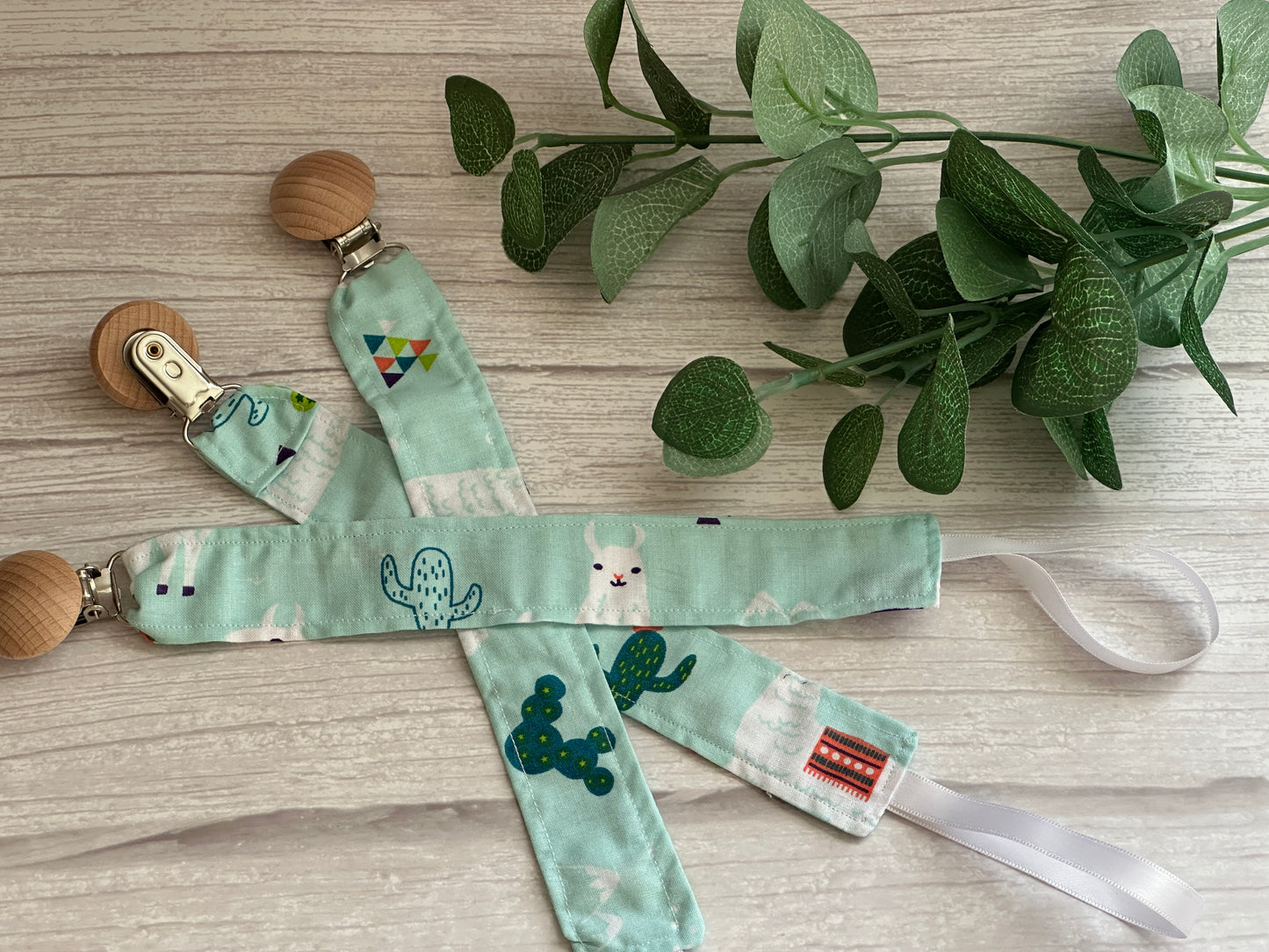 Three pacifier clips from Crafts by Kate's Llama Loves Cotton & Terry Towelling Baby Set, featuring green and cactus-llama patterns, rest on a light wood surface. A small plant with green leaves complements the scene in the background.