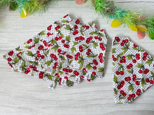 Four triangular pieces of Crafts by Kate's Cotton Pet Bandana with collar attachment - Cherry Sweet are arranged next to one folded piece on a light wood surface, surrounded by colorful decorative strands and small eggs, creating a cozy vibe.