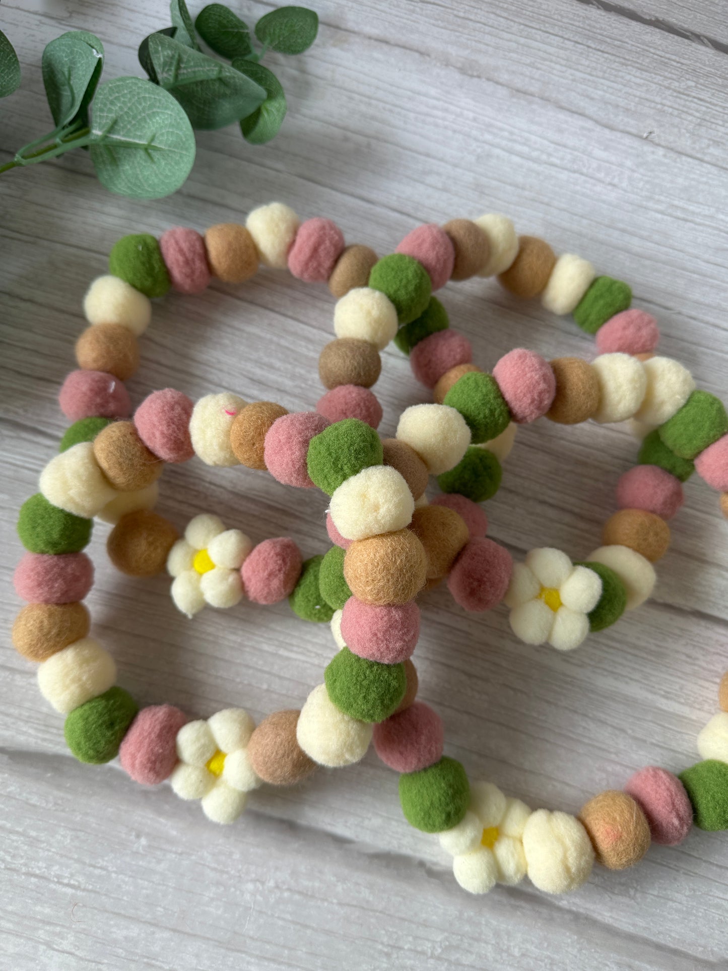 Five PomPom Pet Collar - Fresh as a Daisy garlands by Crafts by Kate, featuring soft cream, pink, green, and brown felt balls with white daisy flowers and yellow centers, are arranged on light wood beside green leaves.