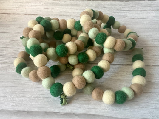 The PomPom Pet Collar - Dino Fun by Crafts by Kate features round felt balls in green, beige, and brown, coiled on light wood, with a playful green felt dinosaur charm at one end.
