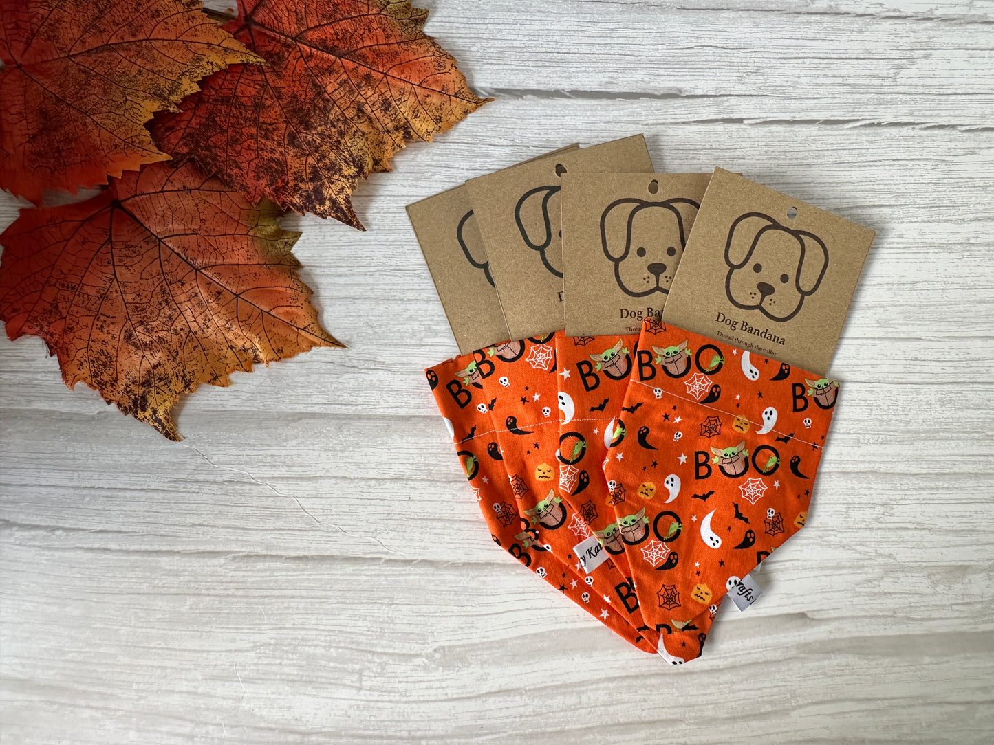 The Crafts by Kate Cotton Pet Bandana with collar attachment - Halloween Baby Yoda is shown on a white wooden surface with autumn leaves. The bandana easily slides onto your pet’s collar and comes in playful, Halloween-themed packaging.